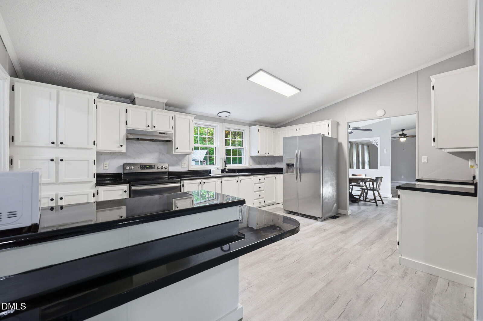 Modern kitchen with white cabinets, black countertops, stainless steel appliances, and light wood flooring. A dining area is visible through an open doorway in the background. Large windows provide natural light.