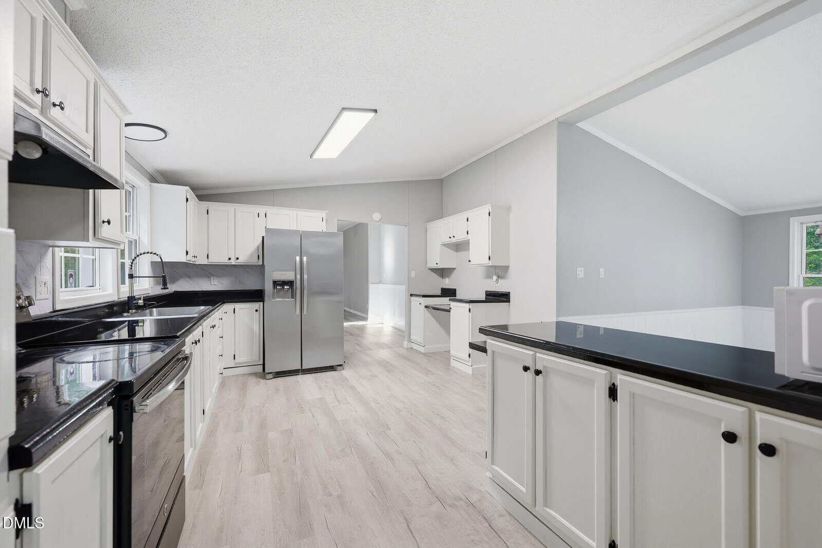Modern kitchen with white cabinets, black countertops, stainless steel appliances, light wood flooring, and an open layout leading to a bright living area with large windows.