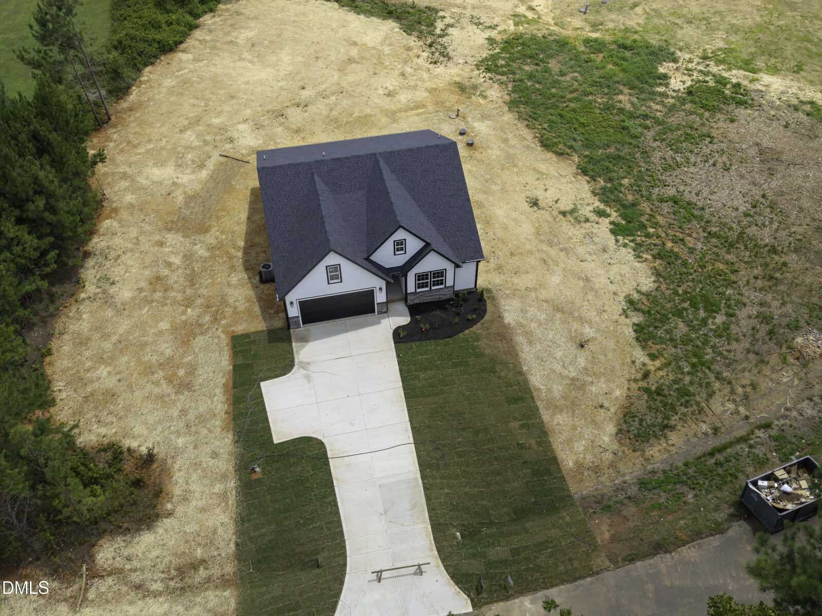 Aerial view of a single-story house with a dark roof, white exterior, attached garage, and new driveway. Sparse grass surrounds the house, with patches of green and brown vegetation nearby.