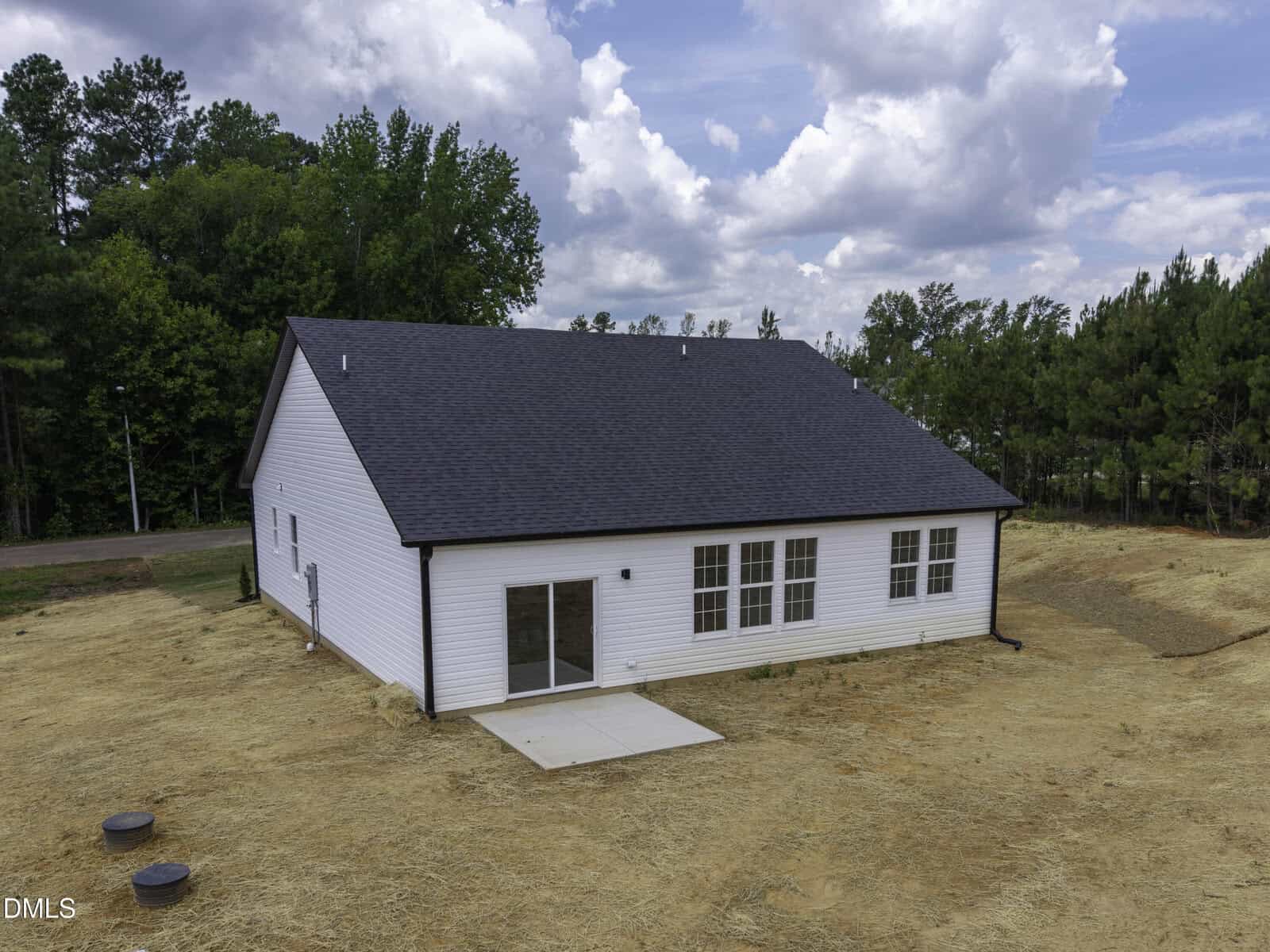 A white single-story house with a dark roof, large windows, and a small concrete patio in a grassy, partially cleared backyard surrounded by trees under a partly cloudy sky.