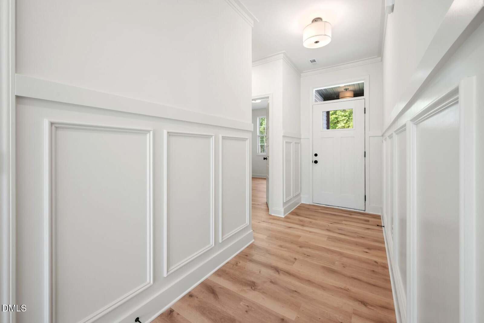 A bright hallway with white paneled walls, light wood flooring, a ceiling light, and a white front door with glass panels. Natural light enters from windows in adjacent rooms.