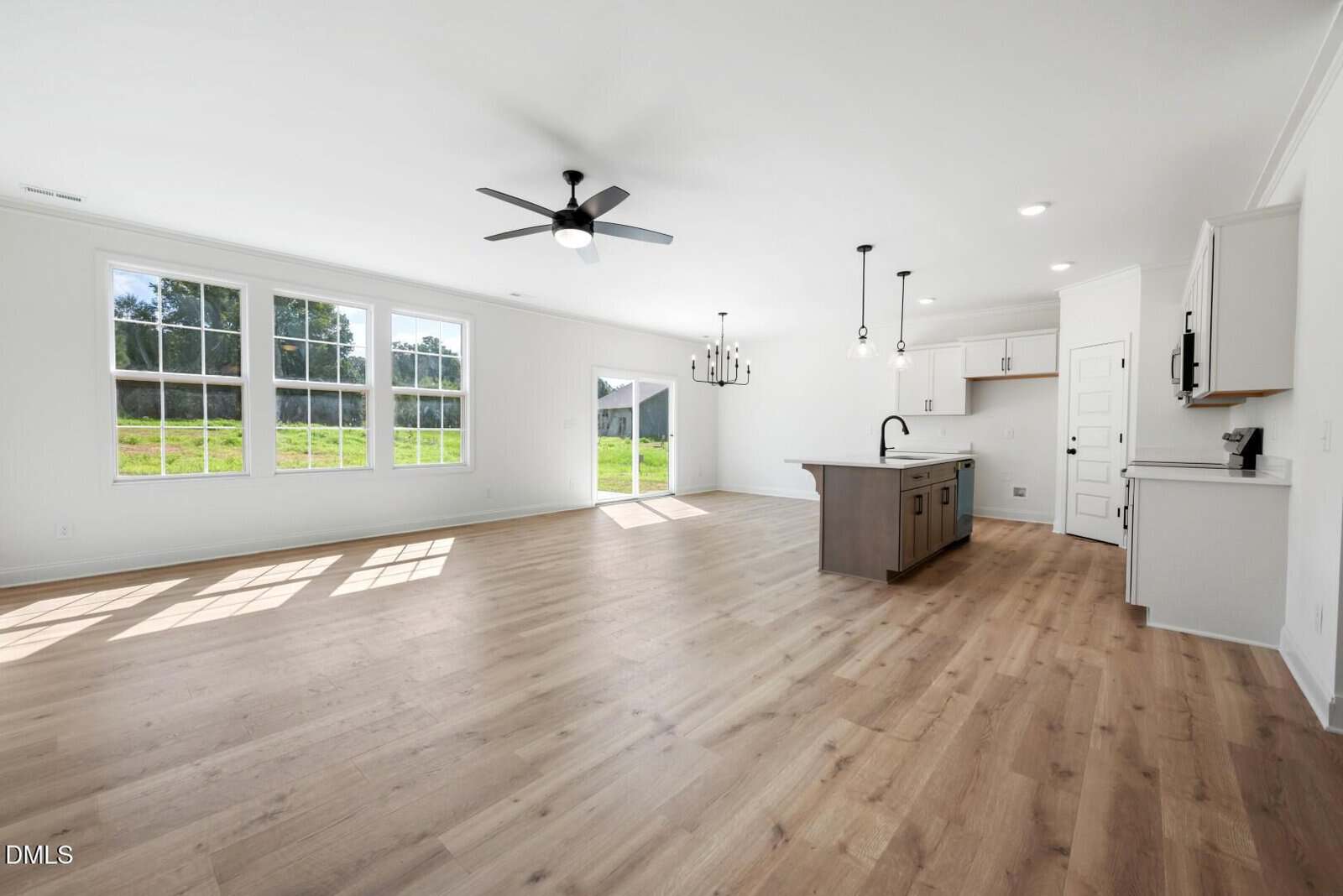 Bright, open-concept living space with large windows, light wood flooring, a ceiling fan, an island kitchen with pendant lights, and a sliding door leading to a grassy backyard.
