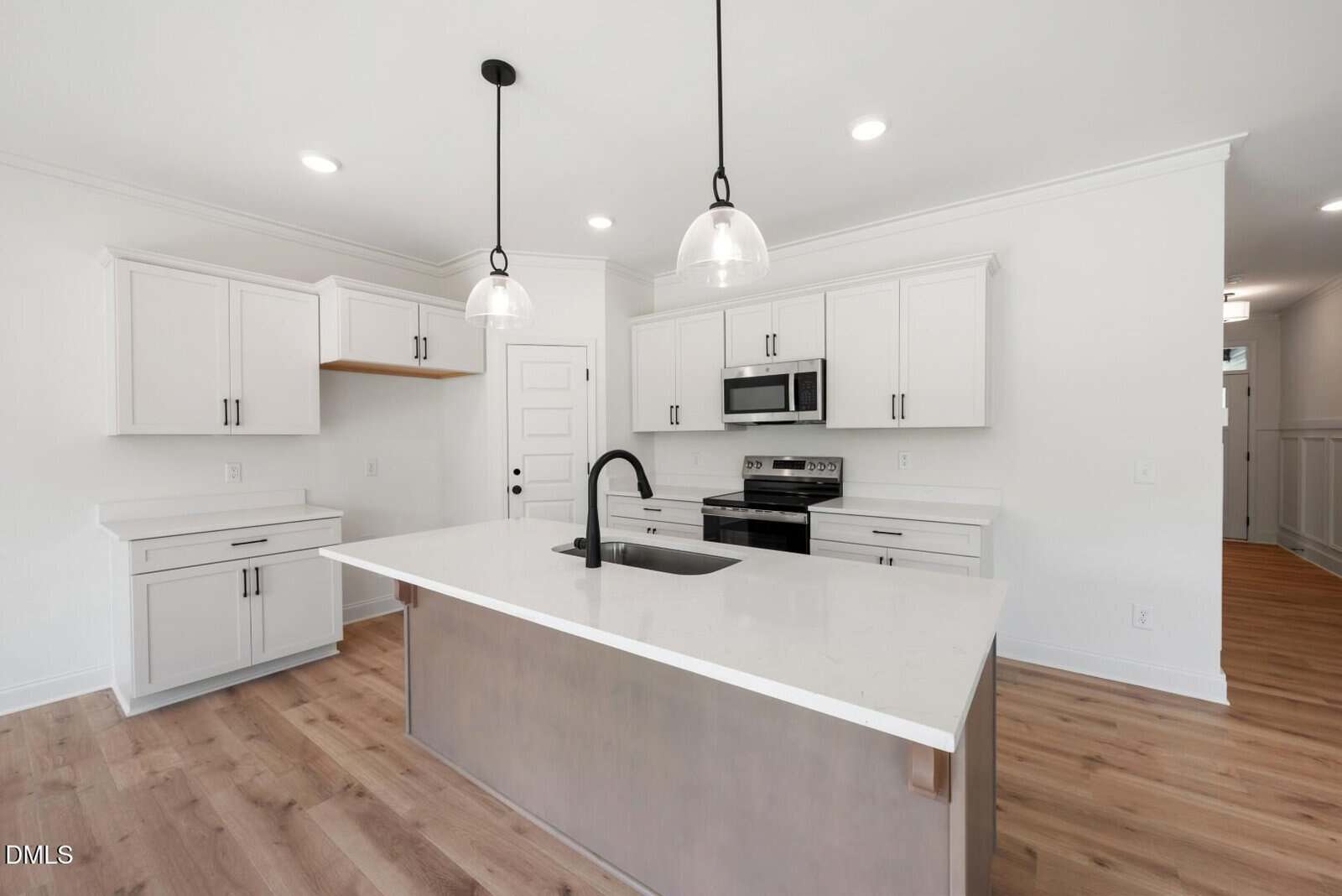 Modern kitchen with white cabinets, stainless steel appliances, an island with a sink and black faucet, two pendant lights, and light wood flooring. A white door and recessed lighting are also visible.