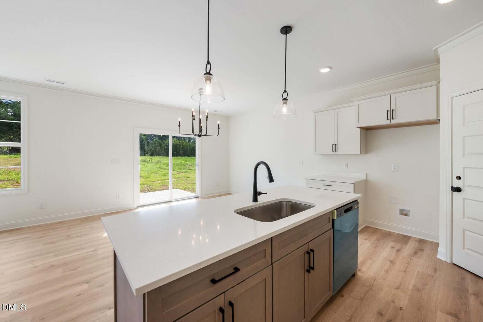 Modern kitchen with a large island, white countertops, dark faucet, built-in sink, dishwasher, and pendant lights. Light wood floors and white cabinets. Sliding glass door leads to a grassy outdoor area.