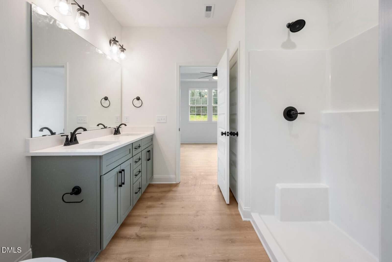 Modern bathroom with double sink vanity, large mirror, three light fixtures, white shower with black fixtures, and light wood floor. An open door leads to a bedroom with a window.