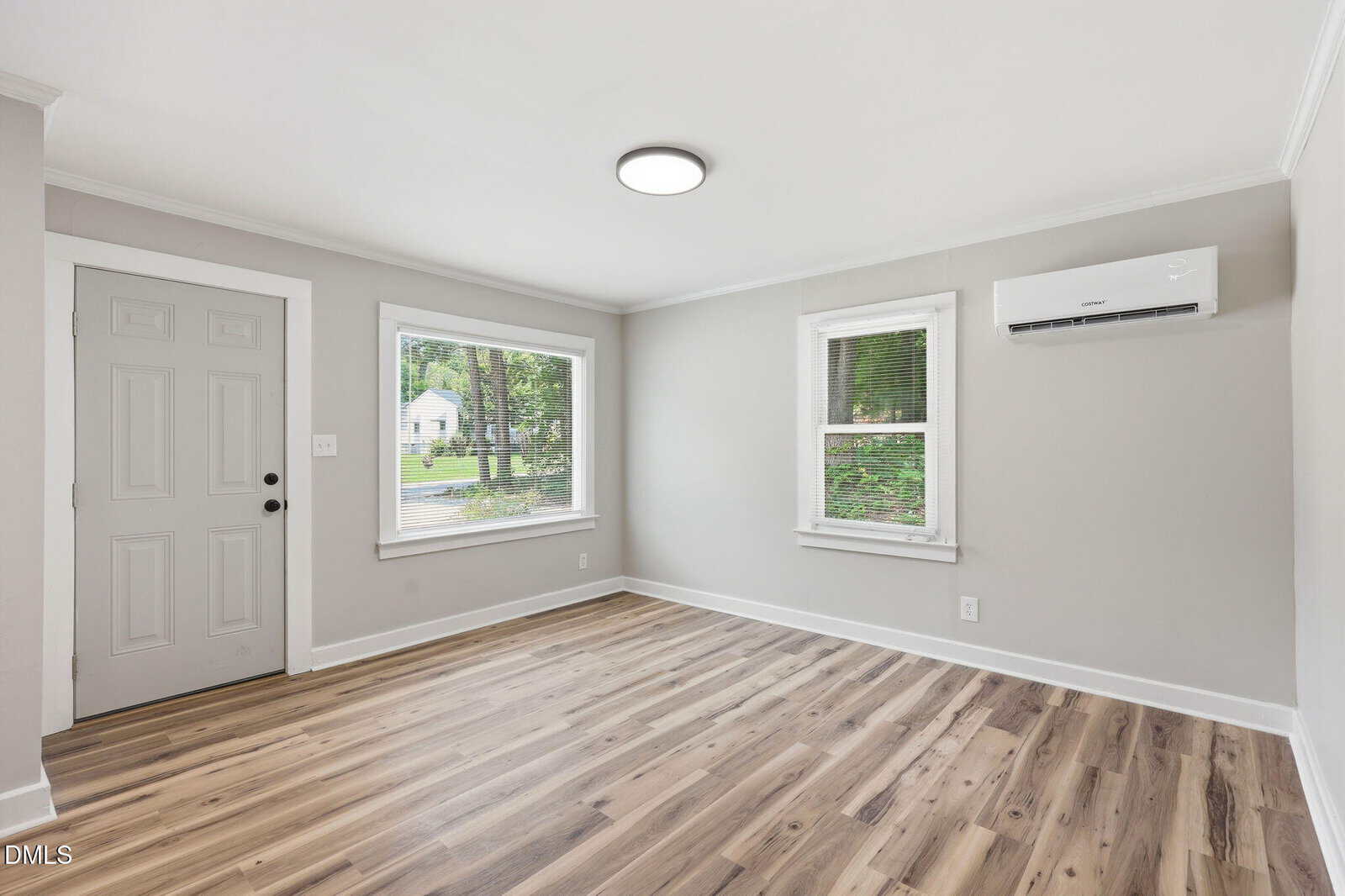 A bright, empty room features light gray walls, wood-style flooring, a window, a door, and a wall-mounted air conditioning unit. Two large windows let in natural light and offer a view of the outdoors.