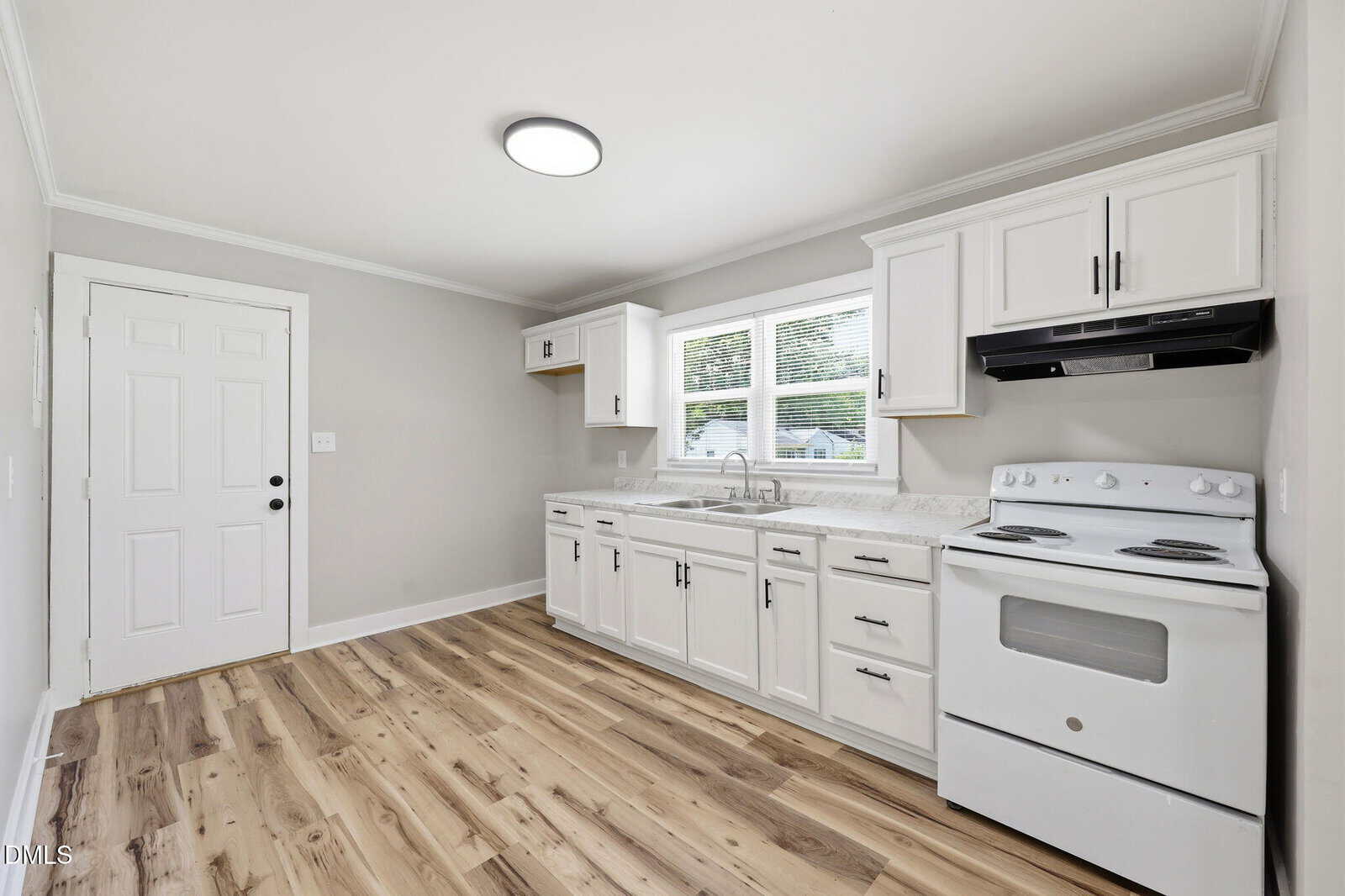 A bright, modern kitchen with white cabinets, a white stove and oven, marble countertops, wood flooring, and a window above the sink. The room has neutral walls and a door leading outside.