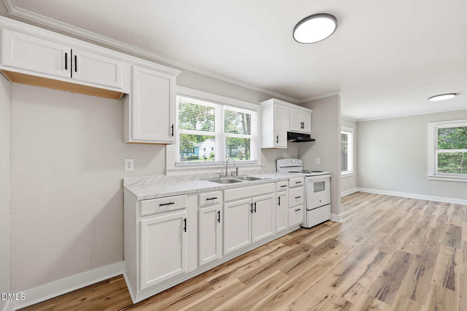 Modern kitchen with white cabinets, marble-patterned countertops, stainless steel sink, white stove, and light wood flooring. Large windows provide natural light, and the adjacent room is visible in the background.