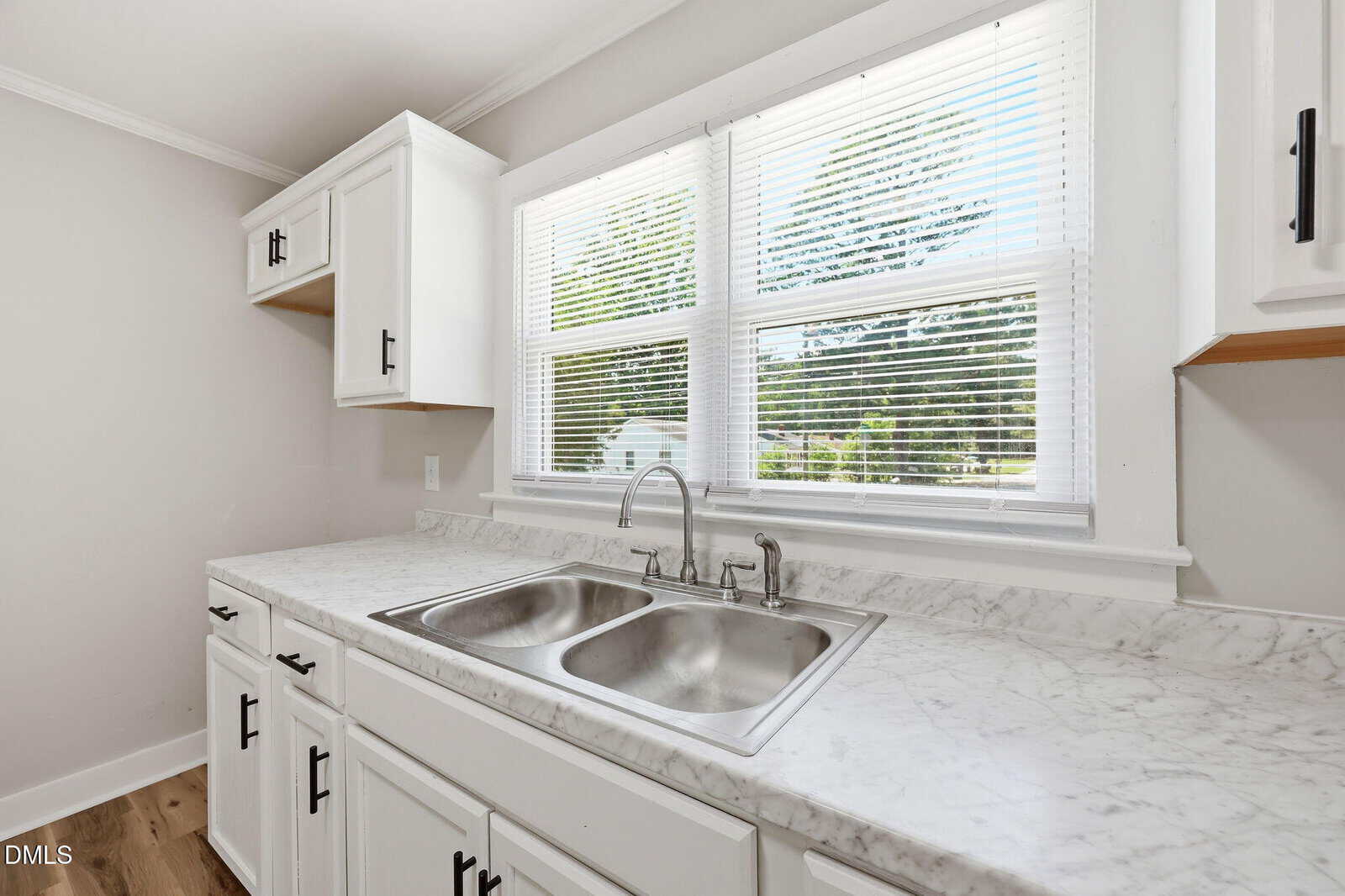 A bright kitchen with white cabinets, a marble-patterned countertop, and a double stainless steel sink beneath a wide window with blinds, letting in natural light.