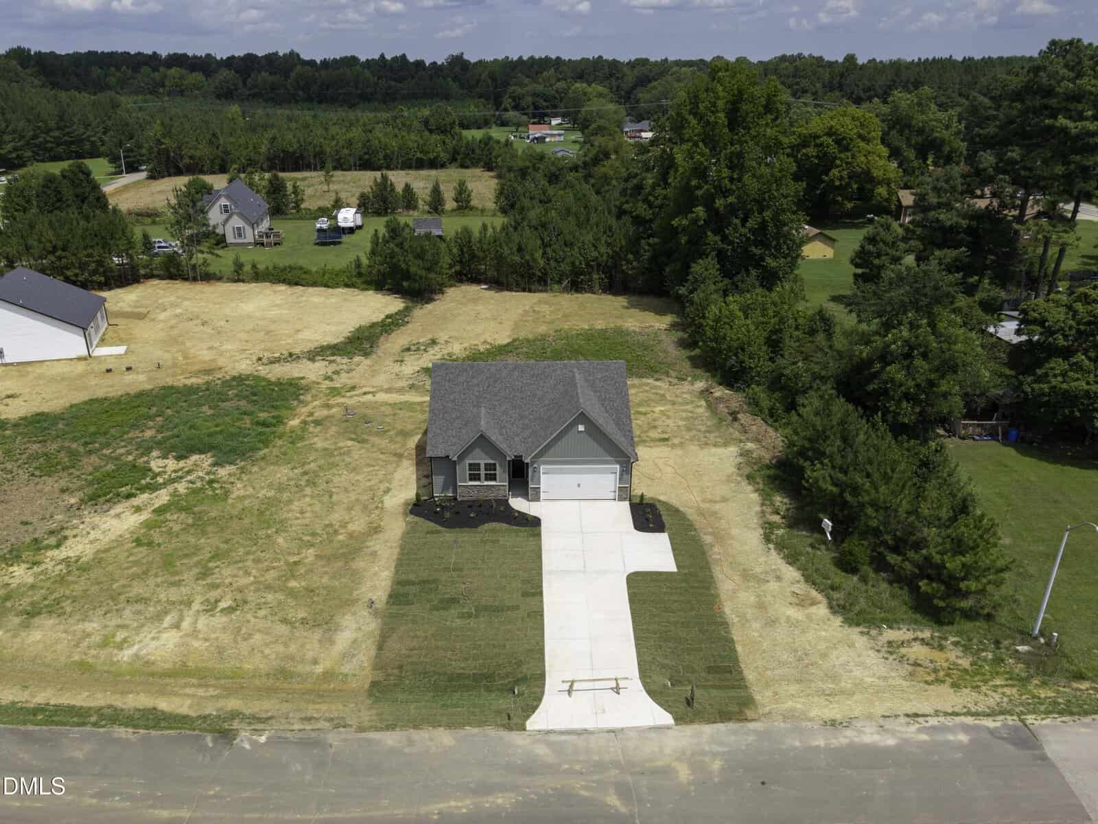 Aerial view of a single-story house with a gray roof and driveway, surrounded by a large, mostly bare yard with patches of grass, trees, and neighboring homes in the distance.