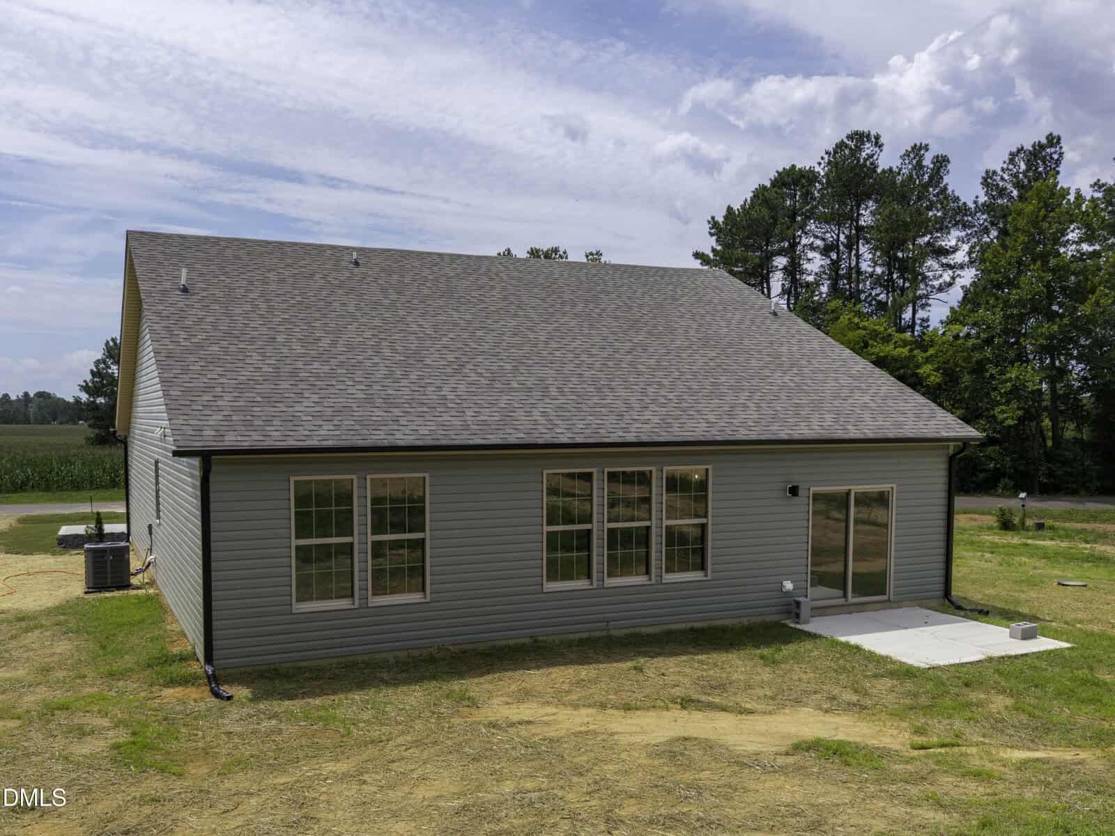 A single-story house with gray siding and a large sloped roof, featuring several windows and a sliding glass door opening to a small concrete patio. Grass and trees surround the home, under a partly cloudy sky.