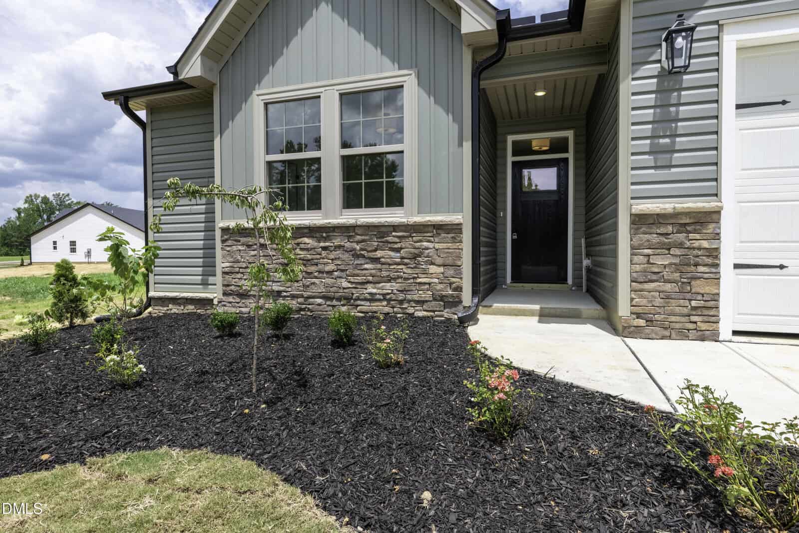 A modern house entrance with light green siding, stone accents, a small garden bed with young plants and mulch, and a concrete pathway leading to the front door under a covered porch.