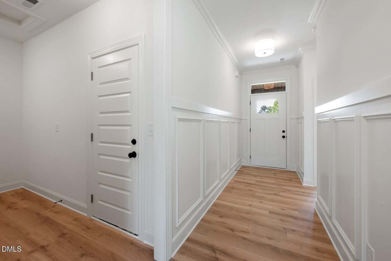Bright hallway with white paneled walls, light wood flooring, white doors with black handles, and a ceiling light near a front door with window panels. Natural light comes through the small transom window above the door.