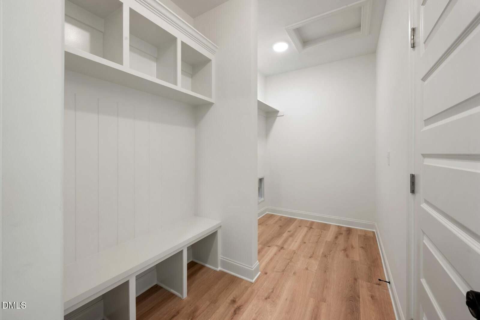 A mudroom with white walls, built-in cubbies and bench, wood flooring, and a closed white door. Ceiling light and attic access visible. Room extends further back.