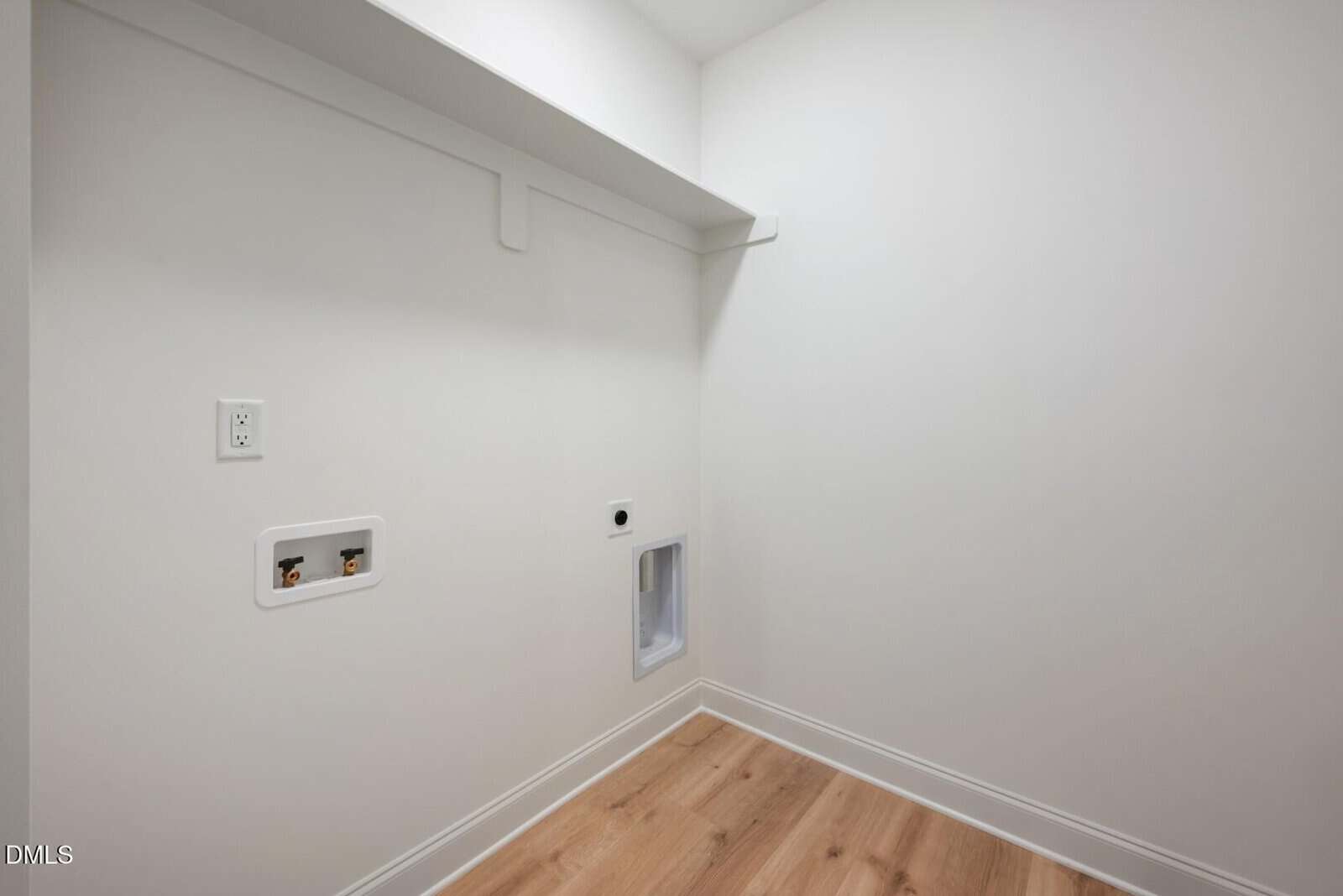 A small, empty laundry room with white walls, wood flooring, washer and dryer hookups, an outlet, and a shelf above the hookups.