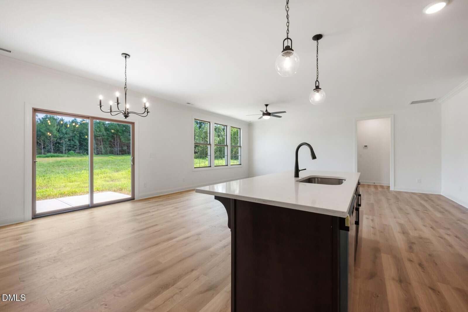 An empty modern kitchen and living area with wood floors, white walls, a large island with sink, pendant lights, a chandelier, and sliding glass doors opening to a grassy backyard.