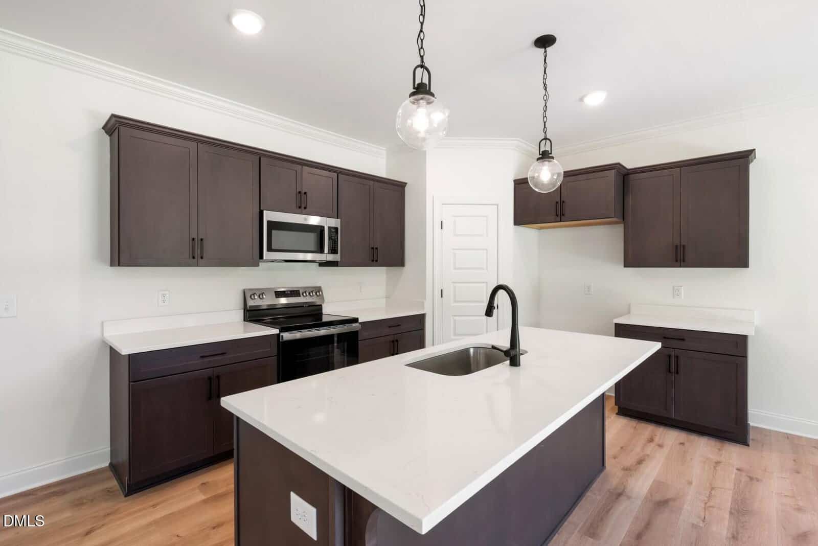 Modern kitchen with dark wood cabinets, stainless steel appliances, white countertops, a central island with a sink, two pendant lights, and light wood flooring. The walls are white and there is a white door in the background.