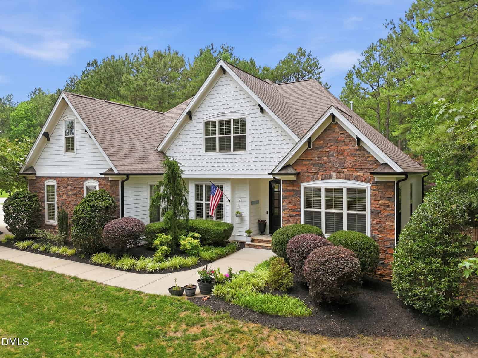 A charming suburban house with a mix of stone and white siding exterior, gabled roof, manicured shrubs, flower beds, and an American flag by the front door, set against a backdrop of tall green trees.