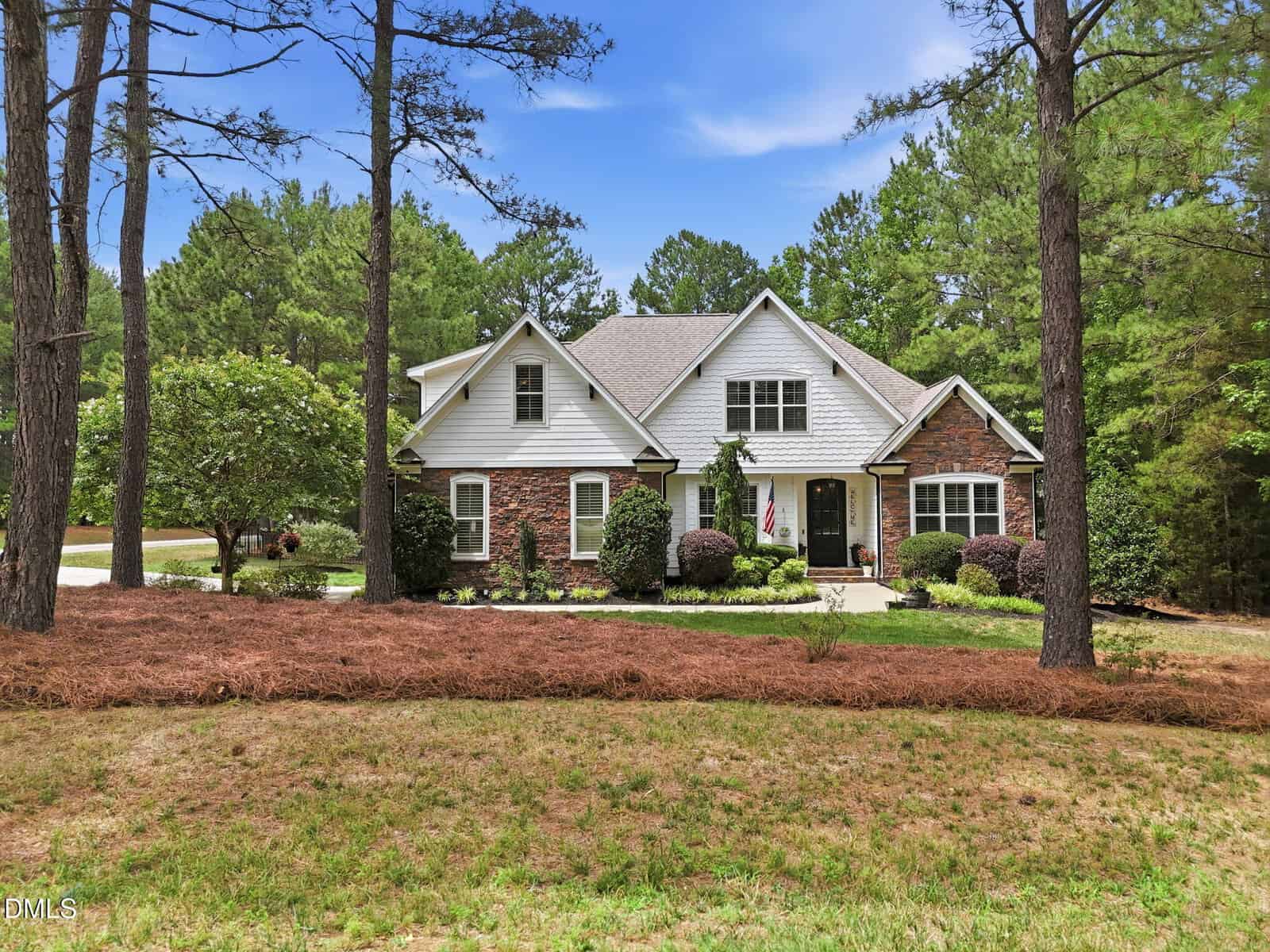 A large house with brick and white siding, surrounded by trees and landscaping, sits behind a lawn with pine straw mulch. An American flag hangs near the front entrance under a partly cloudy sky.