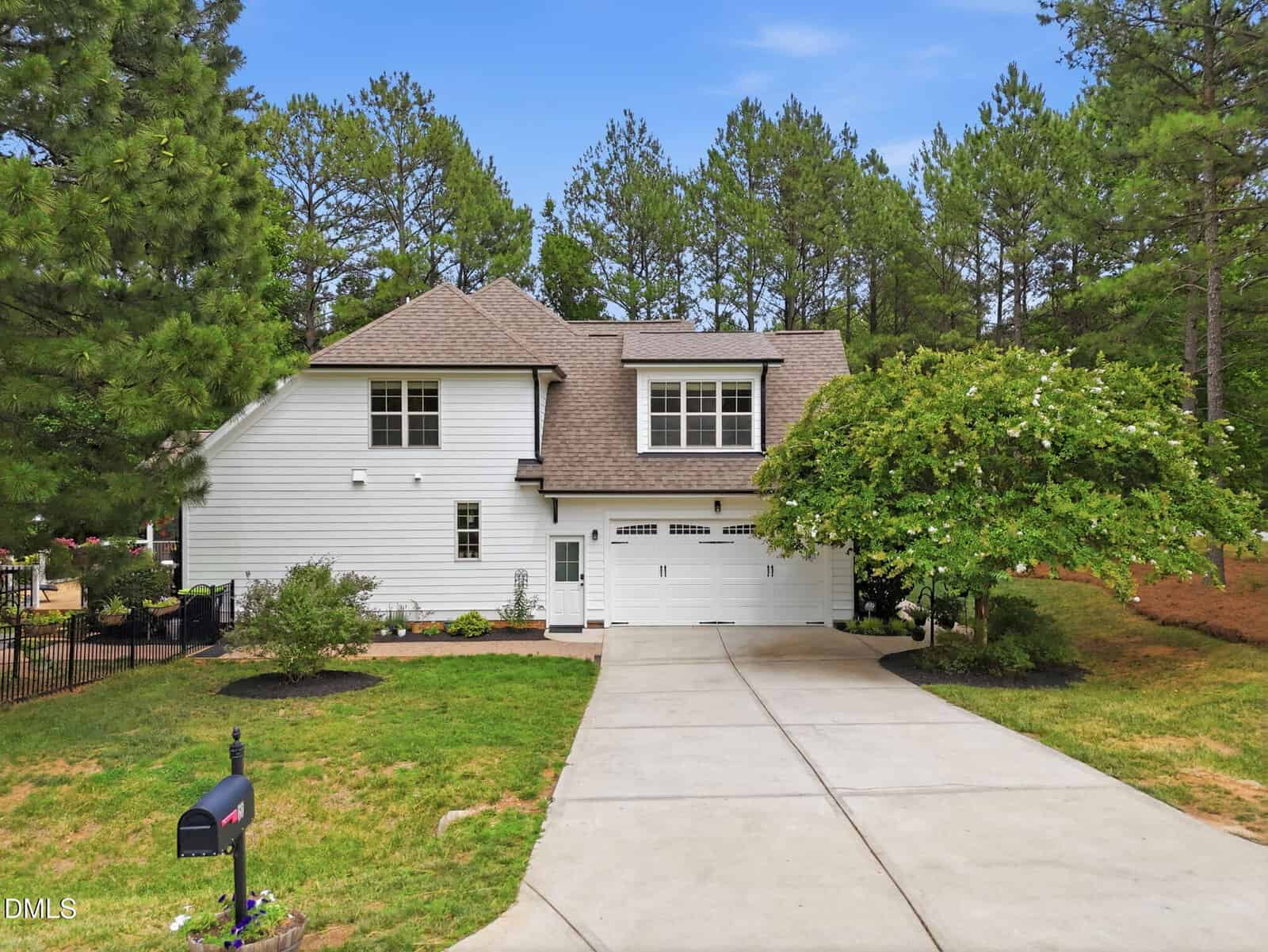 A white two-story house with a double garage, surrounded by trees and greenery, features a wide driveway, manicured landscaping, and a mailbox at the edge of the lawn under a clear blue sky.