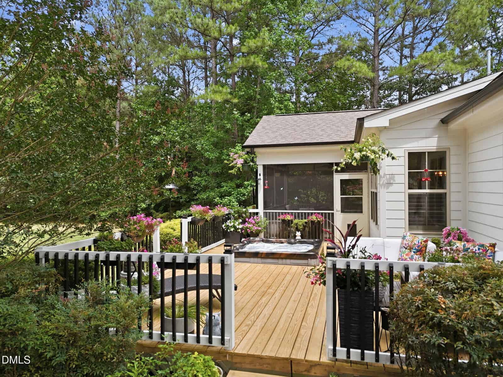 A wooden deck with seating, potted plants, and a hot tub, surrounded by greenery and trees. A white house with large windows and a screened porch is in the background.