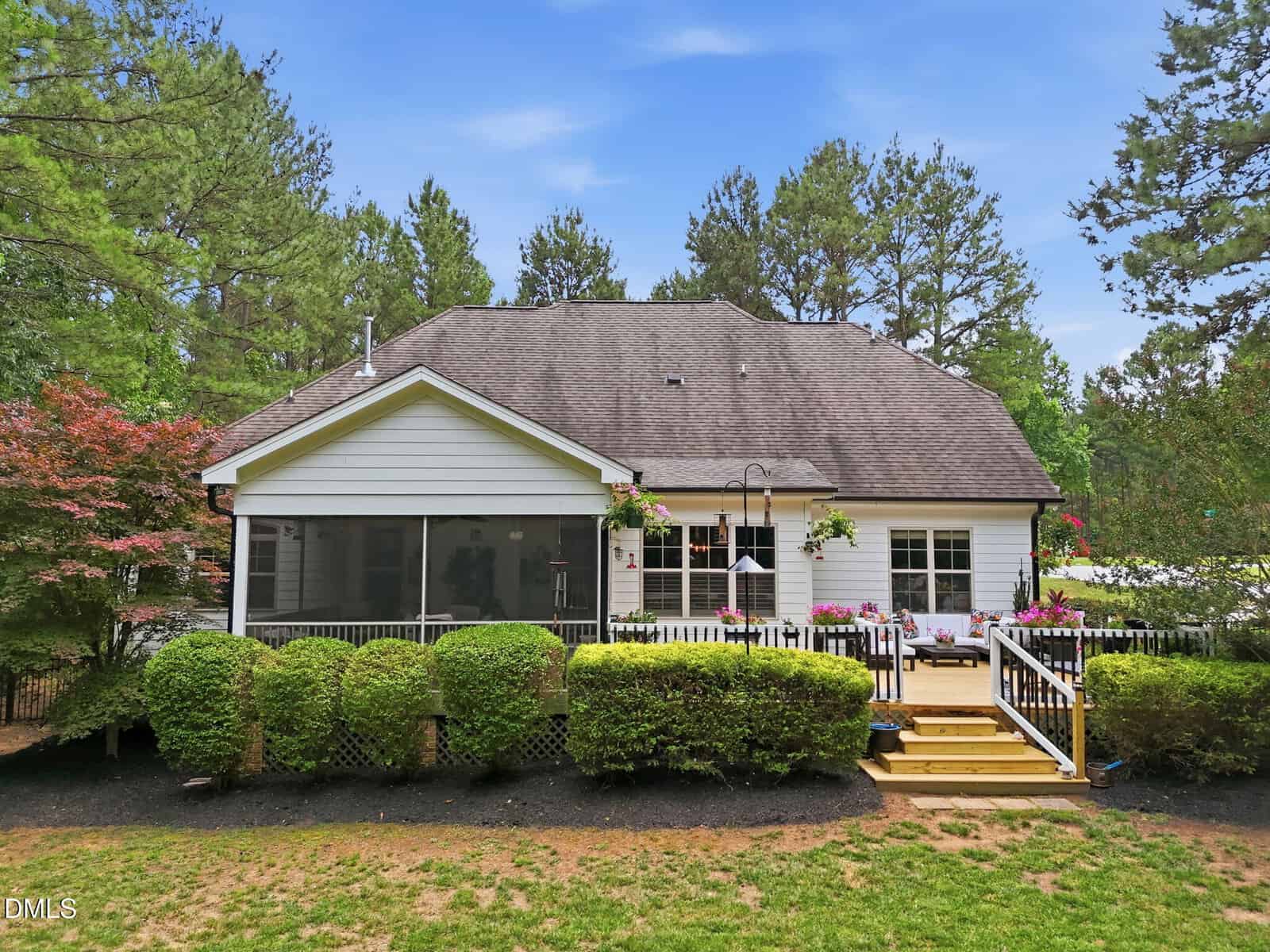 A white house with a gray roof, a screened porch, and a deck with steps. The yard has green bushes and trees surrounding the house under a blue sky.