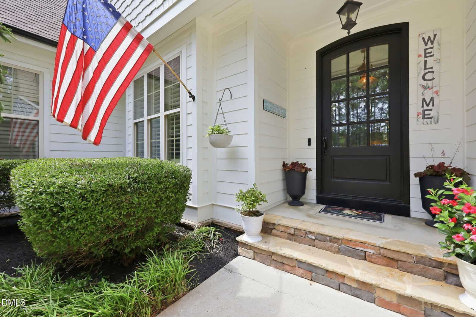 Front porch of a white house with a black door, a "Welcome" sign, potted plants, an American flag, and trimmed bushes alongside the entrance.