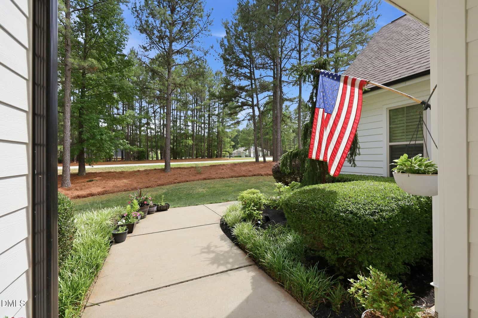 A sidewalk leads past trimmed bushes and potted plants to a yard bordered by tall pine trees. An American flag hangs from the white house on a sunny day.