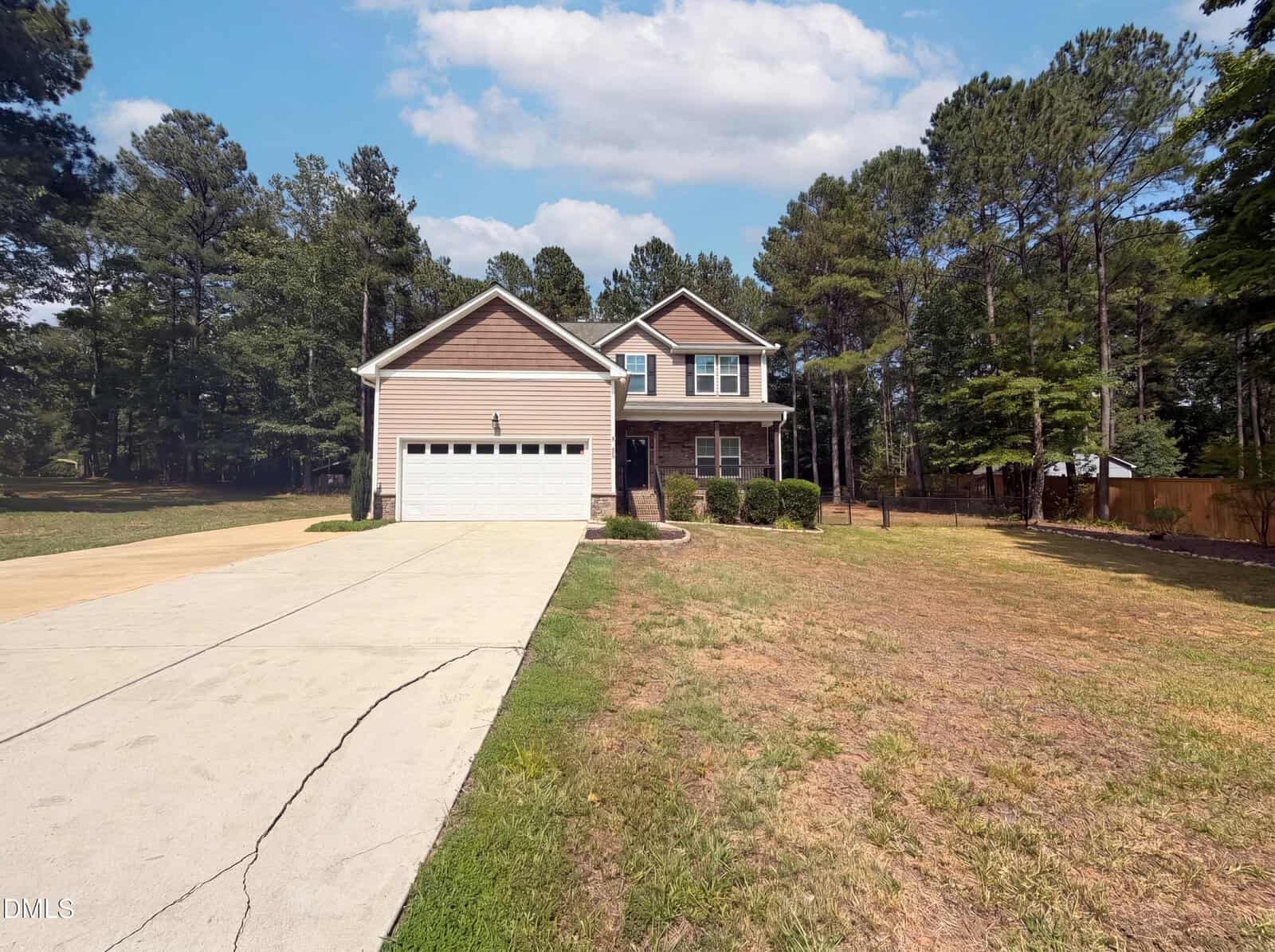 Two-story tan house with attached white garage, wide driveway, and a large front yard with sparse grass. Surrounded by tall trees under a partly cloudy sky.