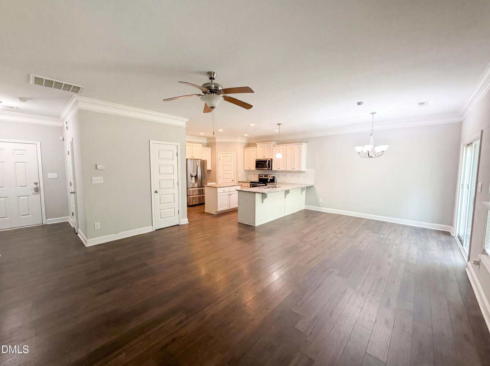 Spacious open-concept living area with dark wood floors, white walls, a ceiling fan, and recessed lighting. The kitchen features white cabinets, a center island, and stainless steel appliances. Large windows let in natural light.
