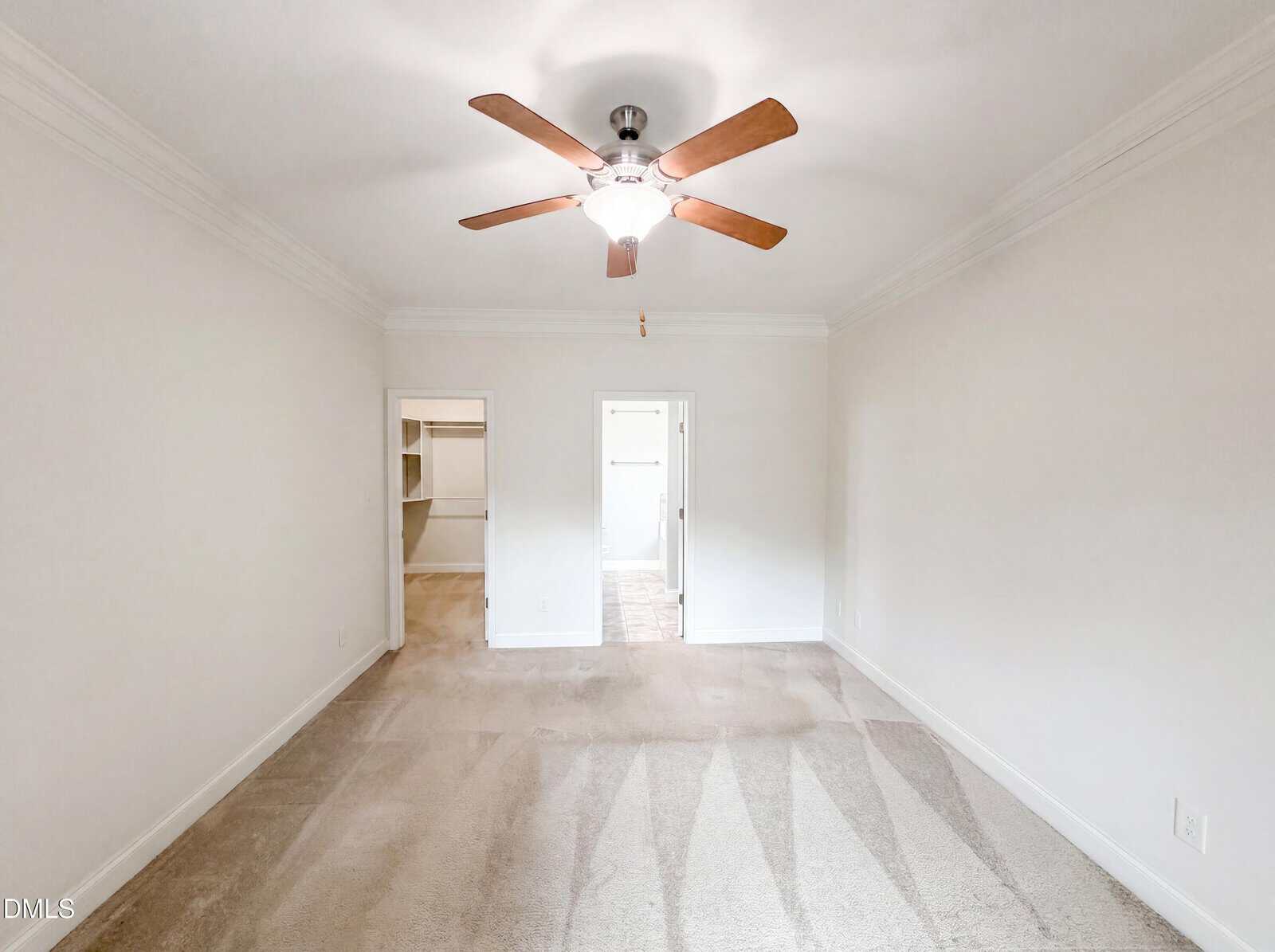 Empty, carpeted room with white walls and ceiling, a ceiling fan with wooden blades, and two open doorways leading to a closet and another room. Bright lighting and neutral colors create a clean, spacious look.