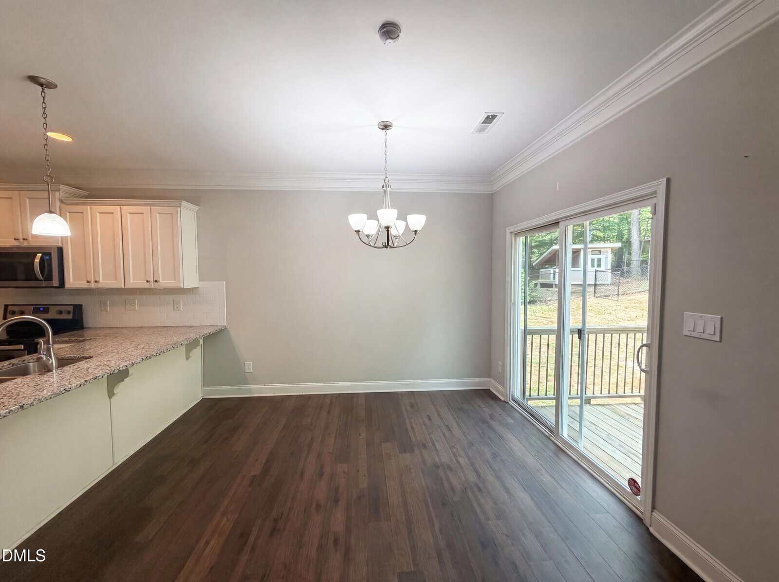 Open kitchen and dining area with dark wood floors, white cabinets, granite countertop, chandelier, and large sliding glass doors leading to a deck overlooking a grassy backyard with trees.