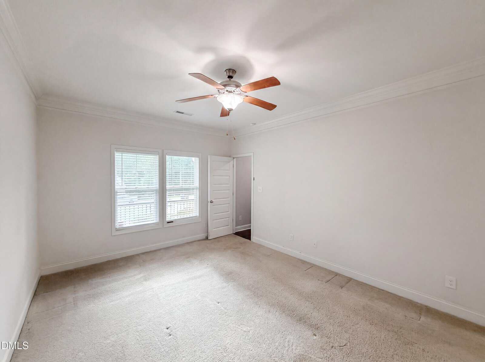 Empty, carpeted bedroom with white walls, a ceiling fan with wooden blades, two windows with blinds, and a white door near the corner. The room is well-lit with natural light.