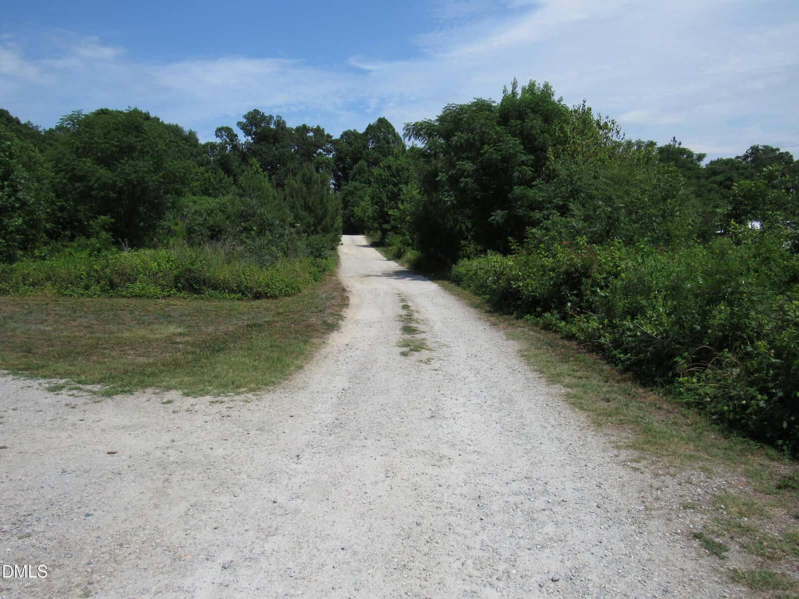 A gravel road stretches into the distance, surrounded by green bushes and trees under a partly cloudy blue sky. The scene is rural and peaceful, with no people or vehicles visible.