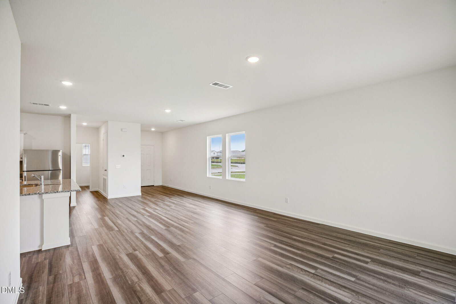 Spacious, empty living area with wood-look floors, white walls, two bright windows, and a view into a modern kitchen with stainless steel appliances in the background.