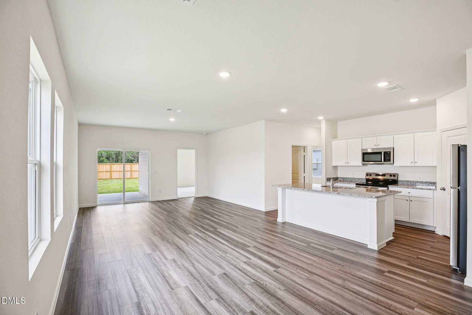 Bright, modern open-concept living area with wood-look flooring, white walls, large windows, a spacious kitchen with white cabinets, stainless steel appliances, and a sliding glass door leading to a fenced backyard.