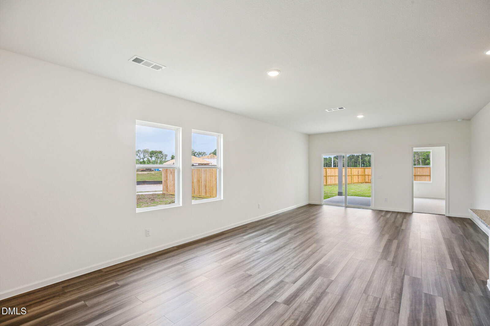 Spacious, empty room with wood flooring, white walls, two large windows, glass sliding doors leading to a fenced backyard, and bright ceiling lights.