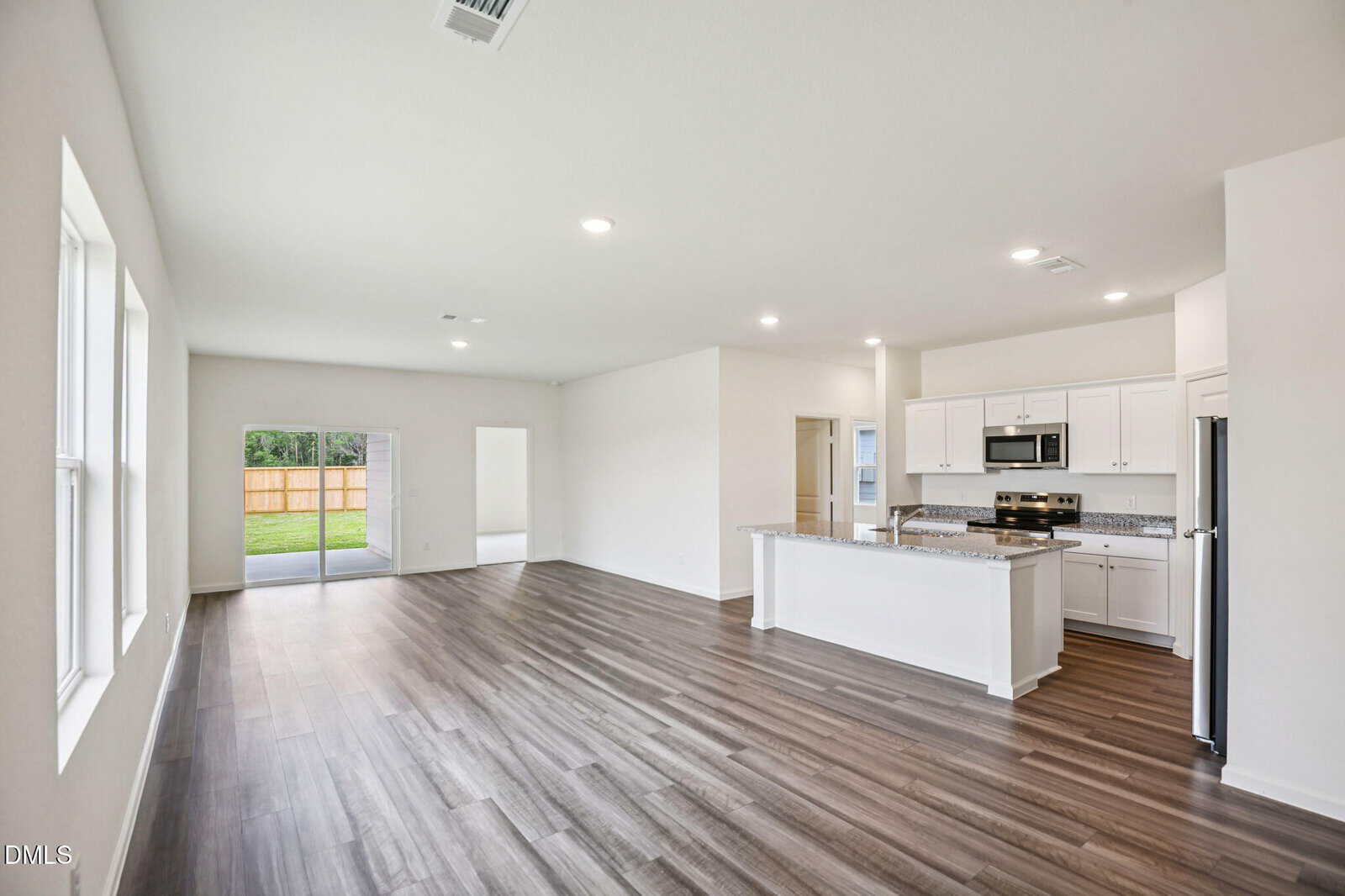 Open-concept living area with wide windows, wood-style flooring, and a modern kitchen featuring white cabinets, granite countertops, and stainless steel appliances. Sliding glass door leads to a fenced backyard.
