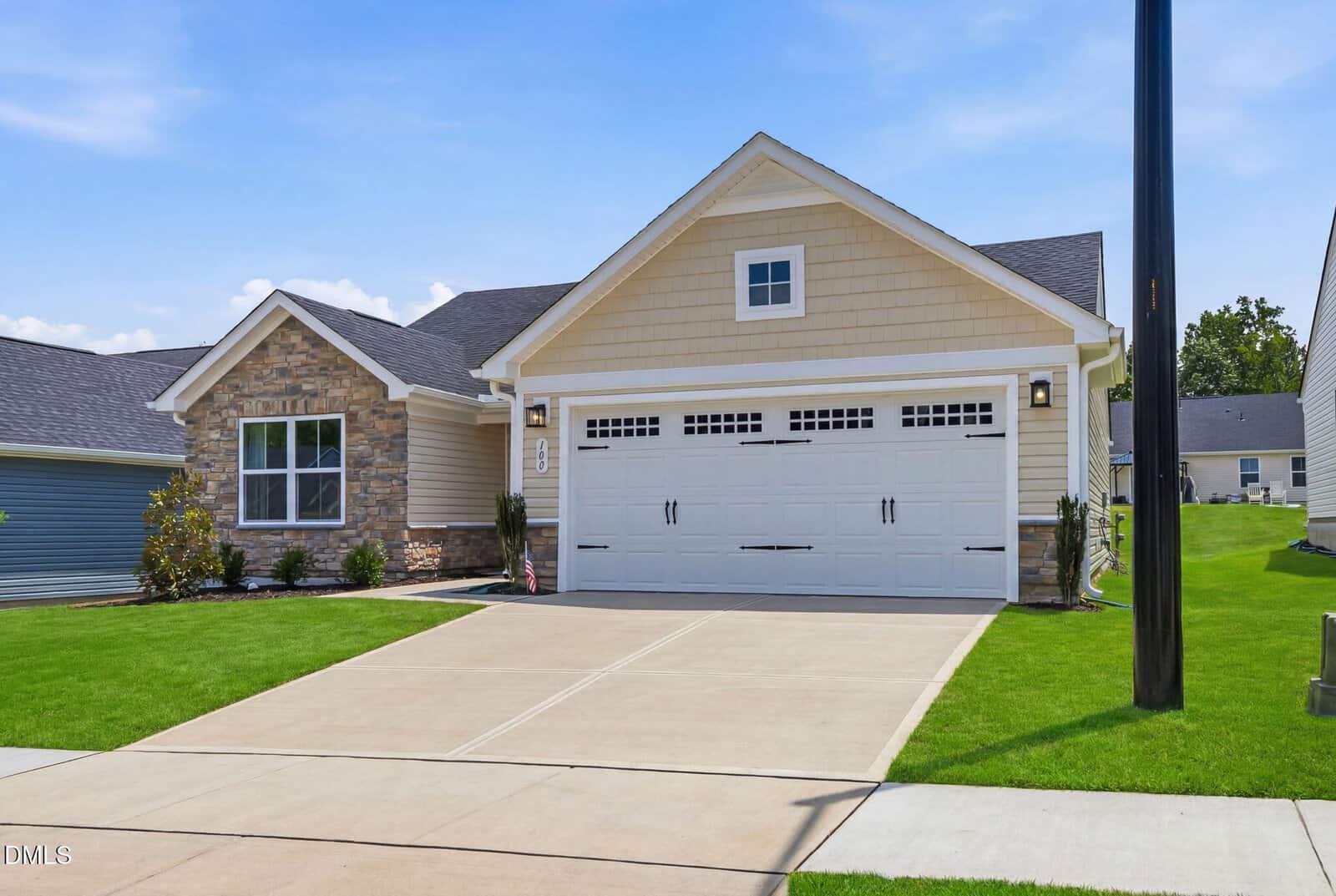 Single-story suburban house with beige siding, stone accents, and a large white double garage door. The home has a well-manicured lawn, wide driveway, and is set on a clear, sunny day.