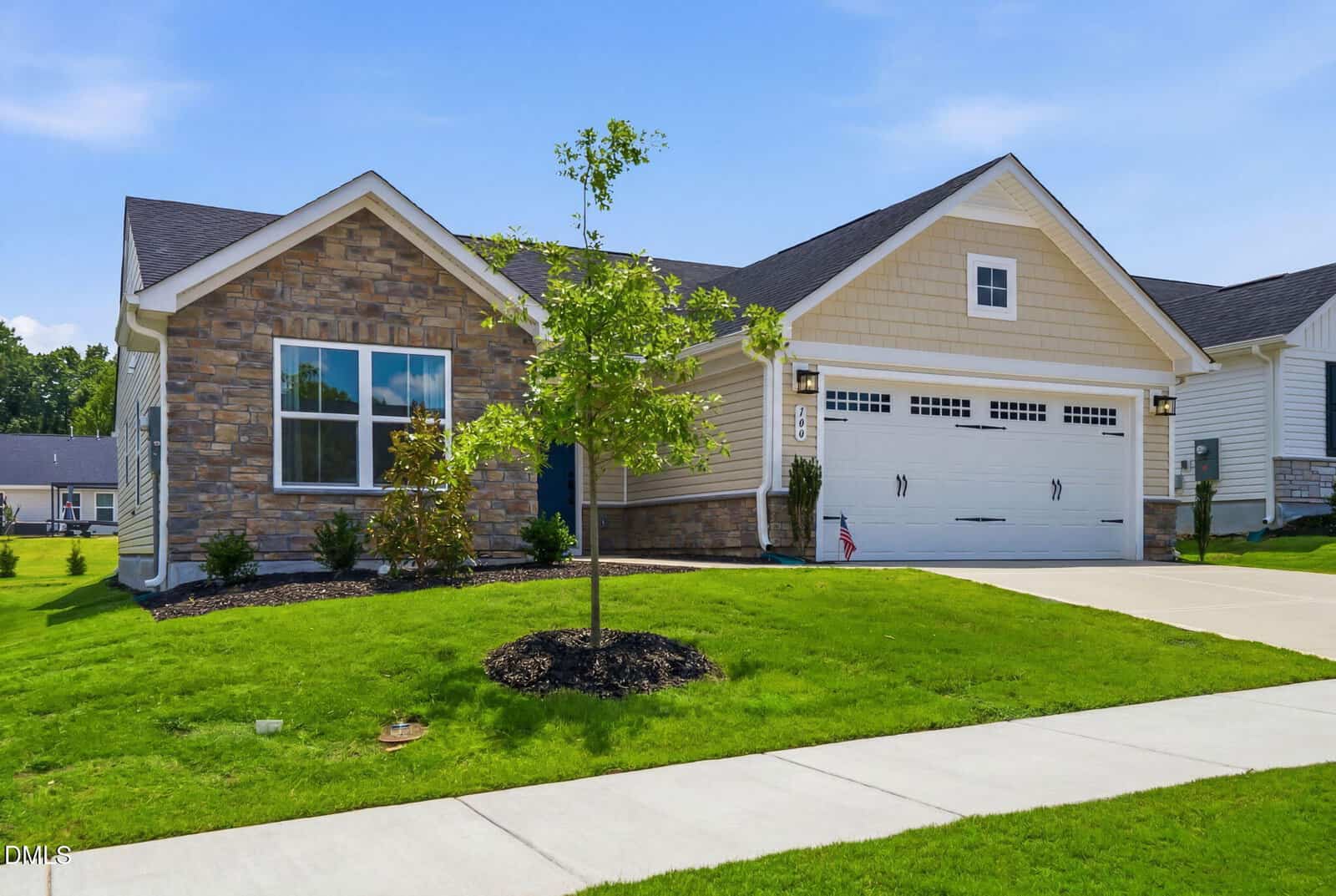 Single-story suburban house with stone and beige siding, a two-car garage with white doors, a small front yard with green grass, young trees, and shrubs, on a sunny day.