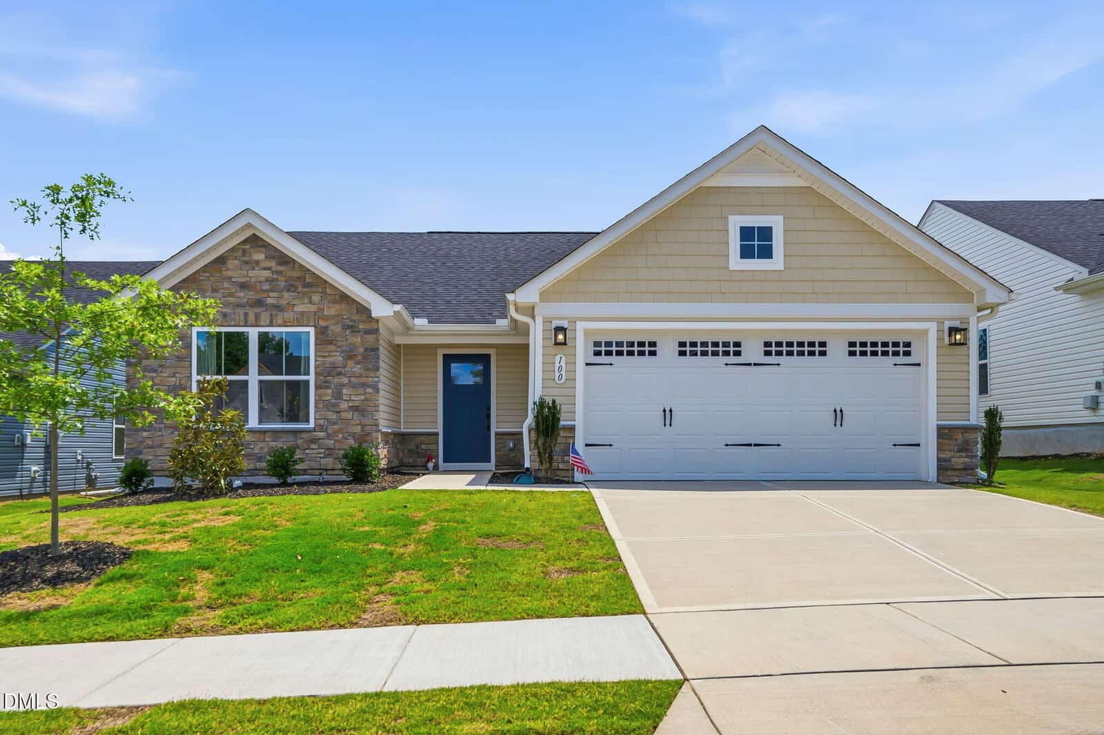 Charming single-story suburban house at 100 Shorebird Lane in Franklinton, featuring beige siding, stone accents, an attached two-car garage with white doors, a neatly trimmed lawn, a small front porch, and a young tree under a clear blue sky.