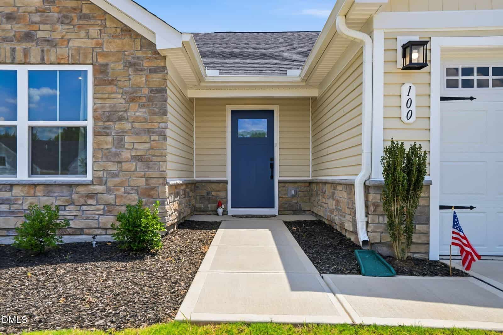 A house entrance with tan siding and stone accents, featuring a blue front door, a small front porch, and a concrete walkway. There are green shrubs and a small American flag near the garage.