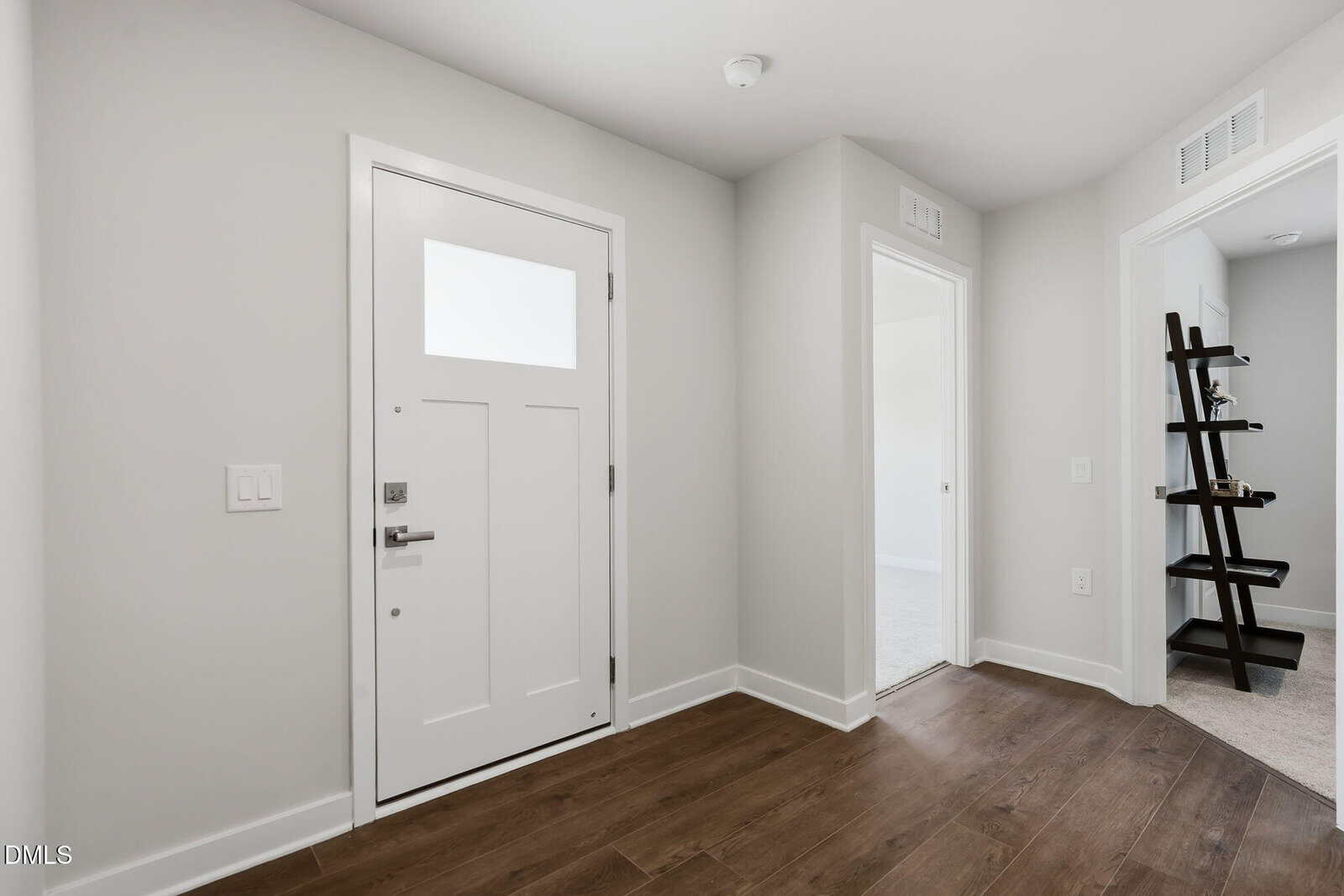 Bright entryway with a white front door, hardwood floors, and light gray walls. Two open doorways lead to carpeted rooms; a dark wooden corner shelf is seen in one room. Space is minimal and modern.