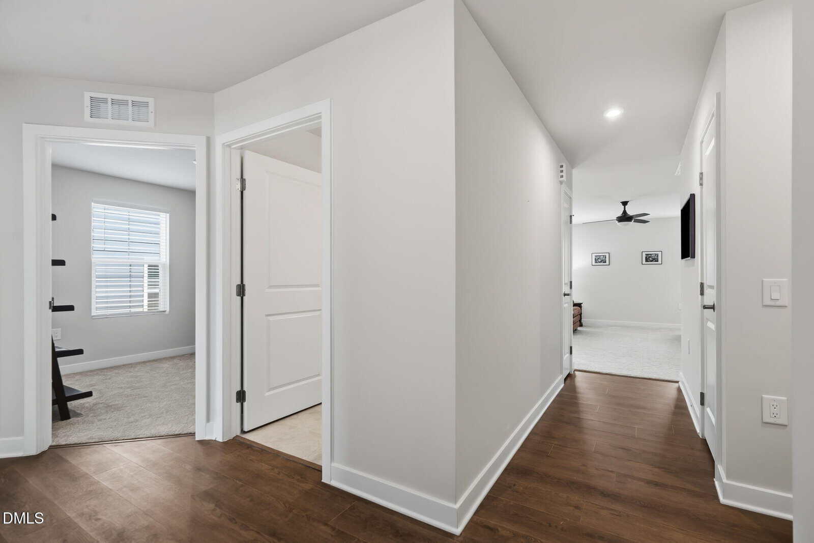 A bright hallway with wood floors leads to carpeted rooms; one door is open, showing a window with blinds and a ladder shelf. Framed photos and a ceiling fan are visible in the distant room.
