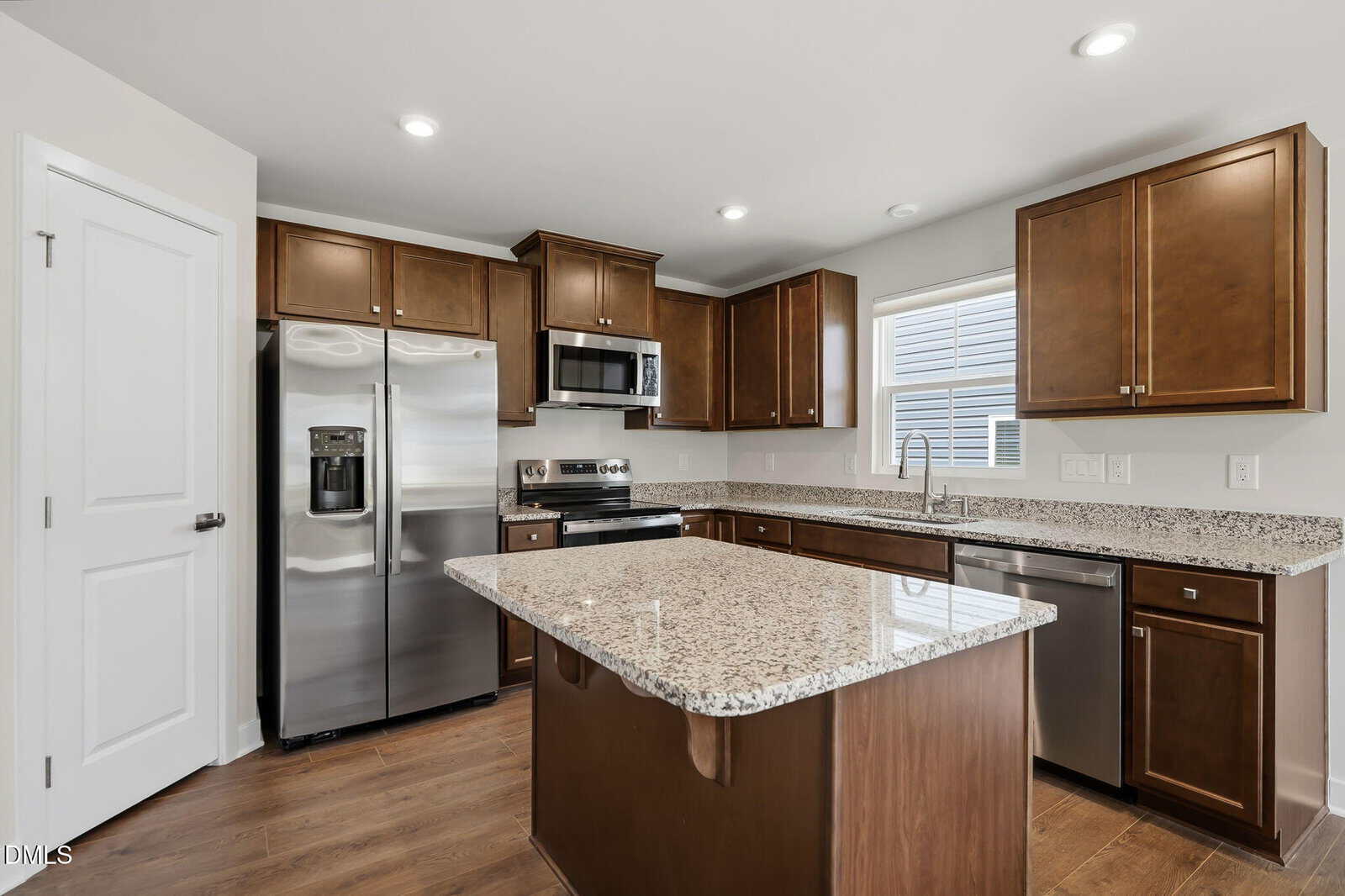 Modern kitchen with brown cabinets, granite countertops, stainless steel appliances, a center island, wood flooring, and a window above the sink letting in natural light.