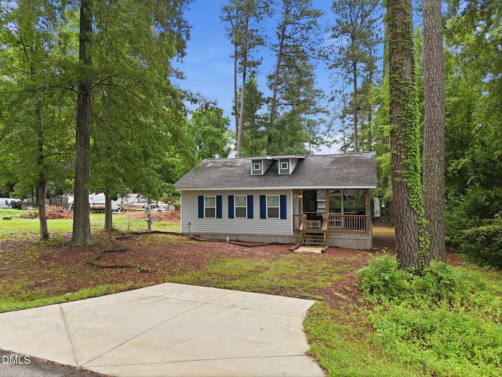 A small, single-story house with beige siding and blue shutters sits among tall trees. The house has a covered front porch and is surrounded by a natural yard with a concrete driveway in front.