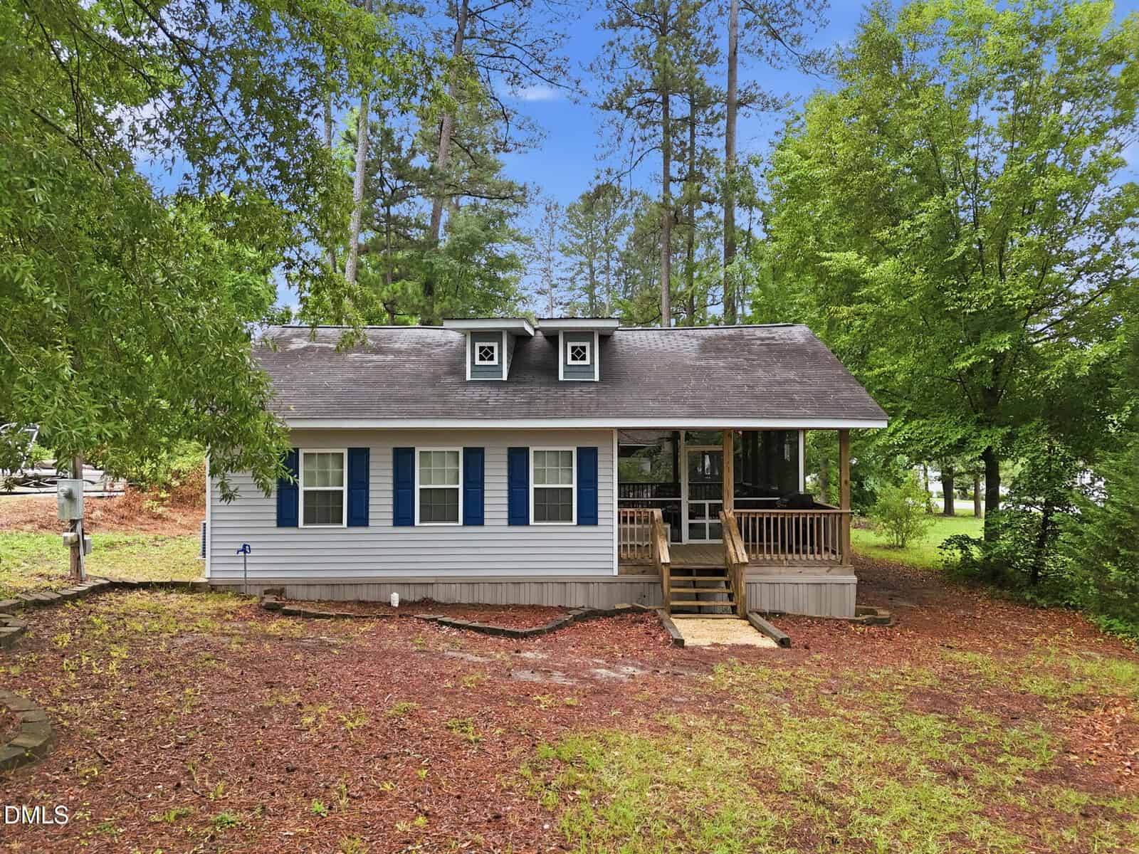 A small, light gray house with blue shutters and a covered front porch sits among tall trees and greenery on a dirt and pine needle-covered yard under a blue sky.