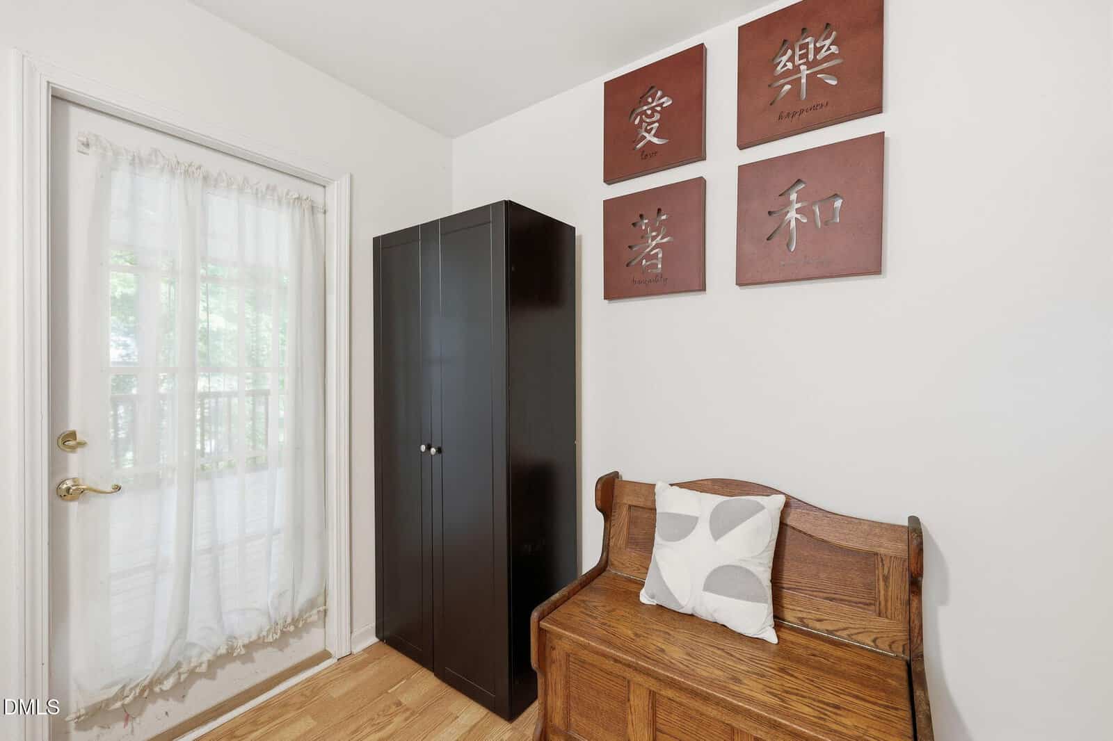 A small room with a glass door with sheer curtains, a dark wooden wardrobe, a wooden bench with a cushion, and four wall art pieces with Chinese characters and English translations.