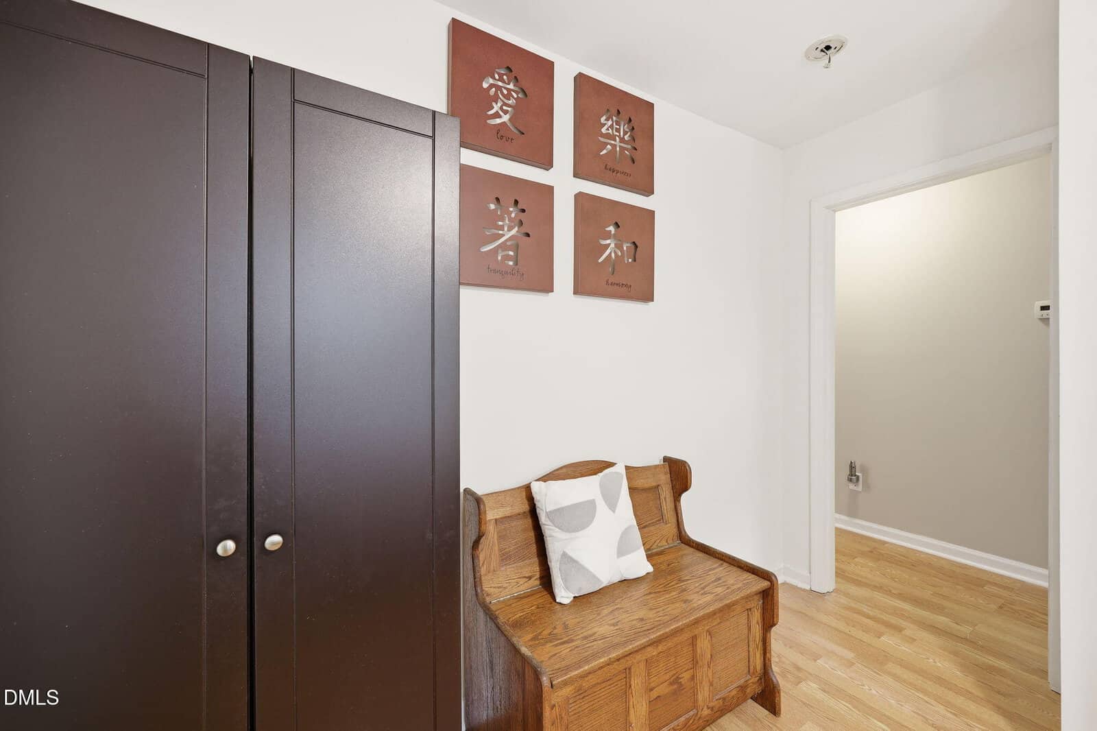 A wooden bench with a pillow sits against a white wall beneath four decorative plaques with Asian characters and words like "Love" and "Harmony." A dark wardrobe is next to the bench on a light wood floor.