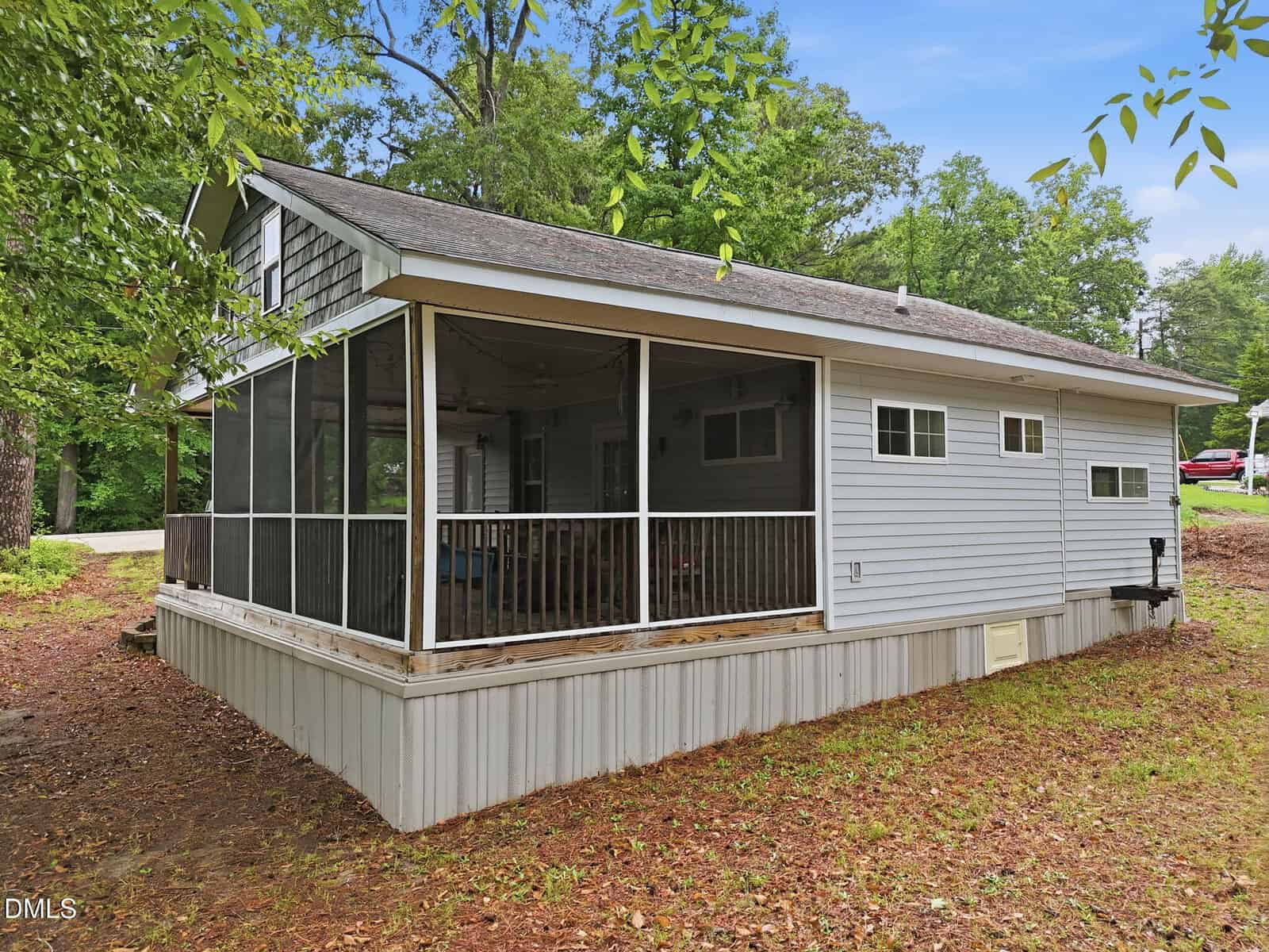 A small light-gray house with white trim features a large screened-in porch and a sloped roof, surrounded by trees and grass under a clear sky.